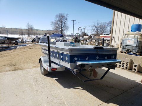 Blue-and-white boat on a trailer in a parking lot, with stars on the side and a folded black ramp.
