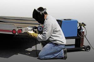 Mechanic kneeling beside a car’s rear bumper, using a tool next to a blue machine on a wheeled cart