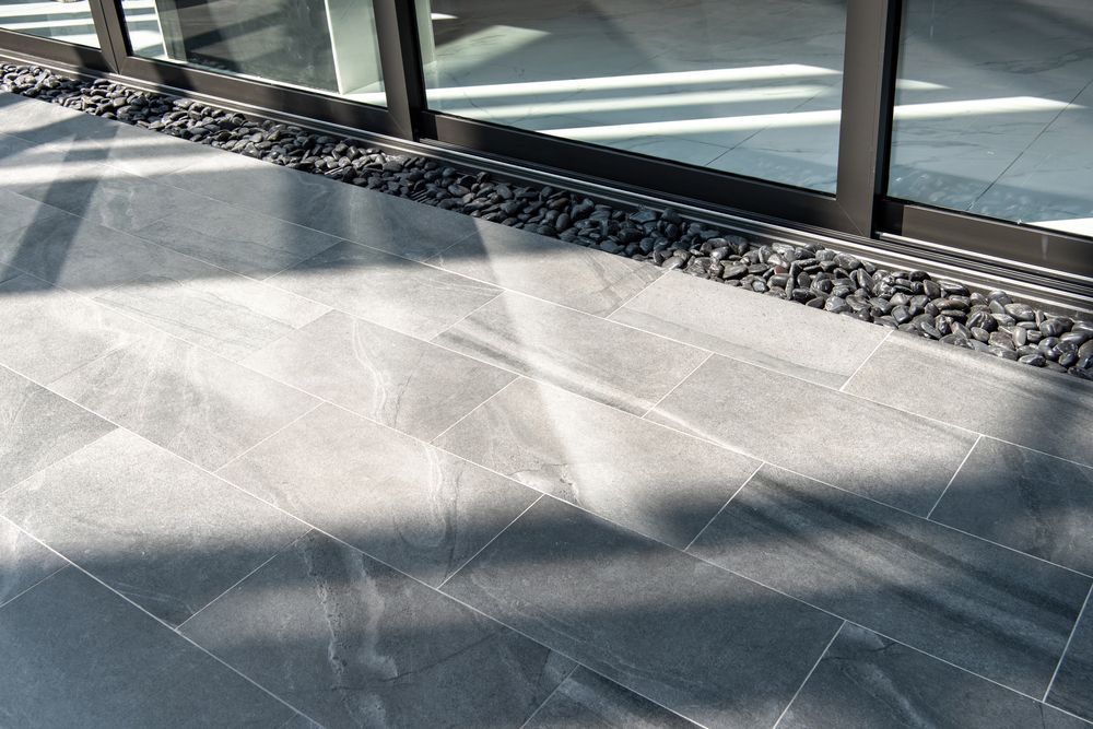 Gray tiled patio with dark pebbles along glass doors, sunlight and shadows.