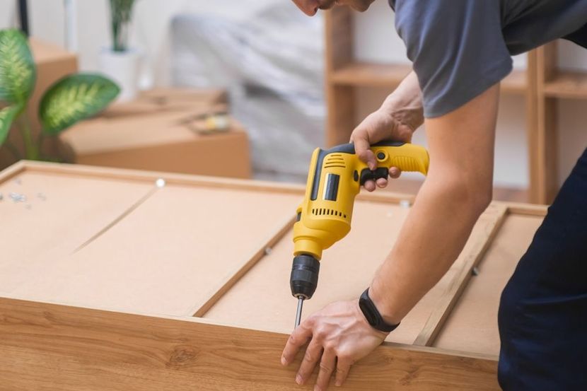 Person using a yellow power drill to assemble furniture. Brown wood and cardboard boxes are in the background.