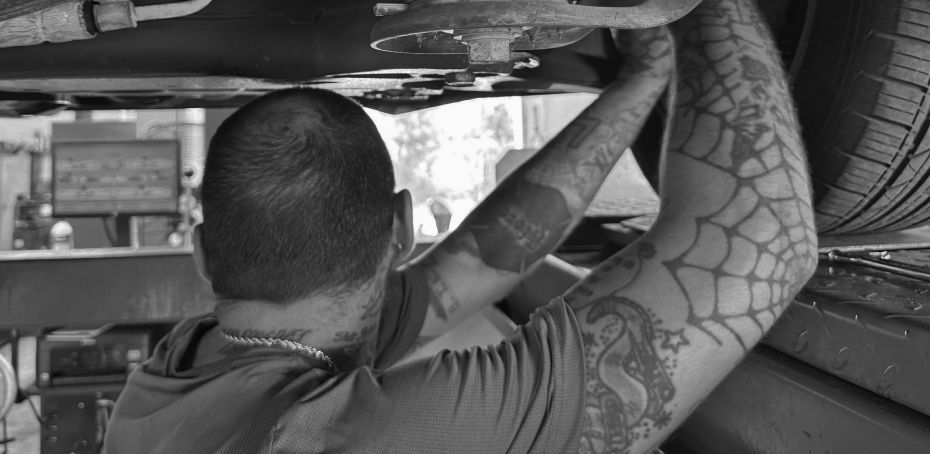 Mechanic working under a vehicle, reaching up with tattooed arms in a garage. | RMP Auto