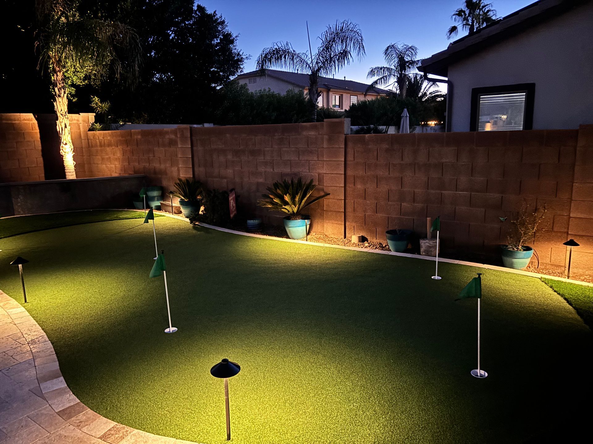 Backyard putting green at night, lit by spotlights, with flags and a brick wall.