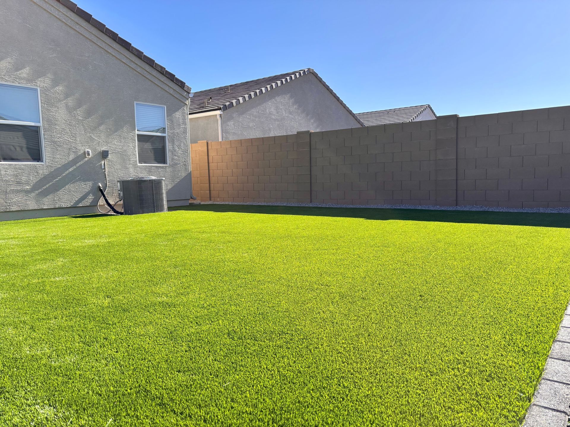 Sunny backyard with green lawn, tan privacy wall, and stucco house under a clear blue sky
