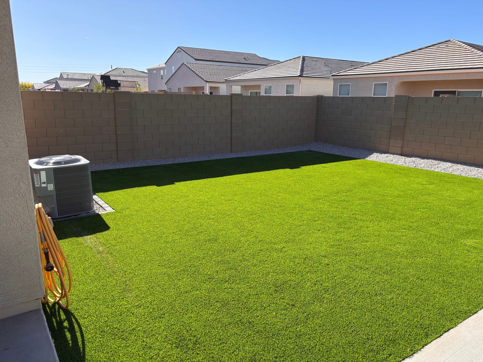 Sunny fenced backyard with artificial turf, gravel border, and AC unit.