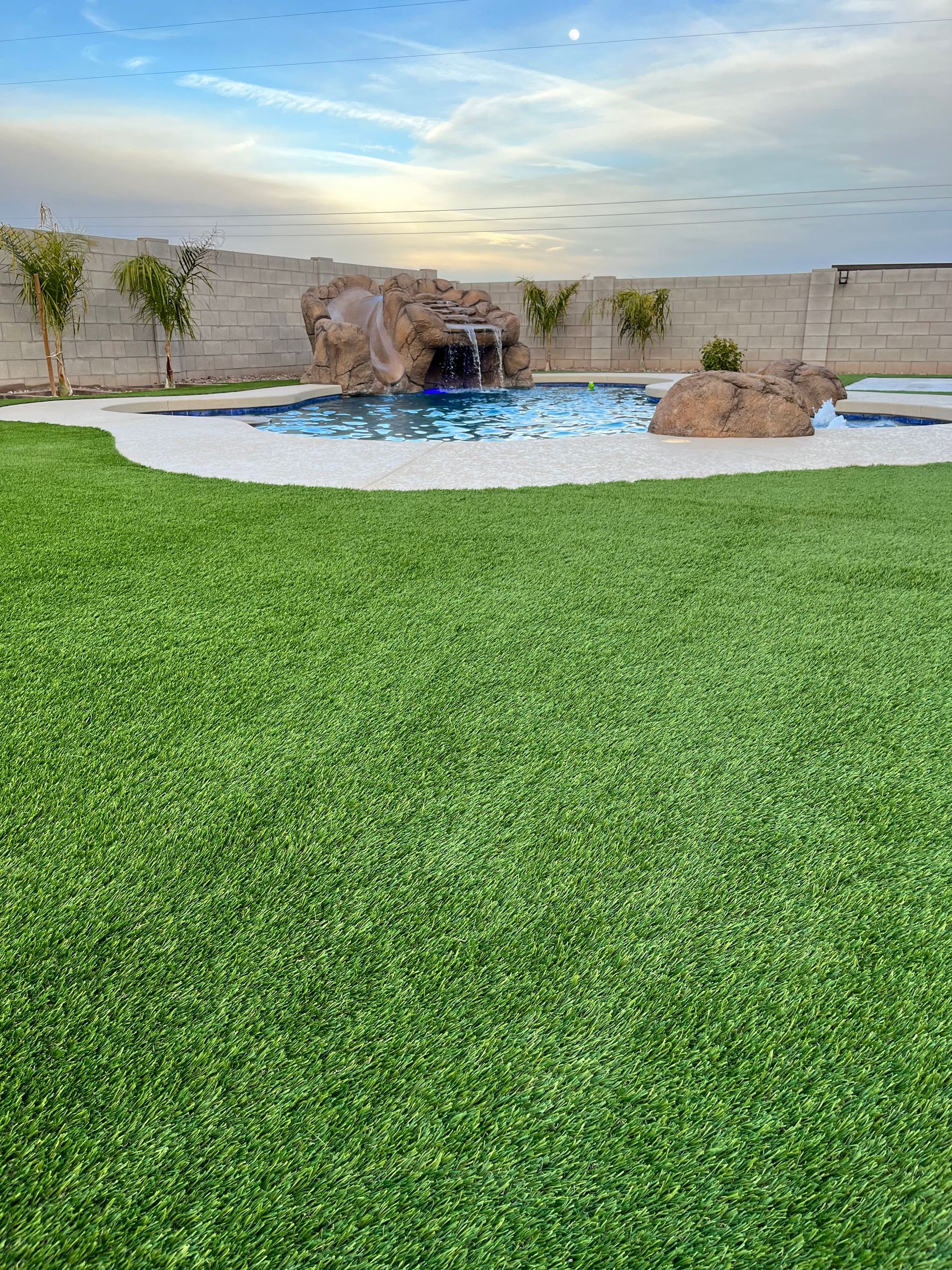 Curved backyard pool beside bright green lawn at sunset, with rocks and a desert-style landscape.