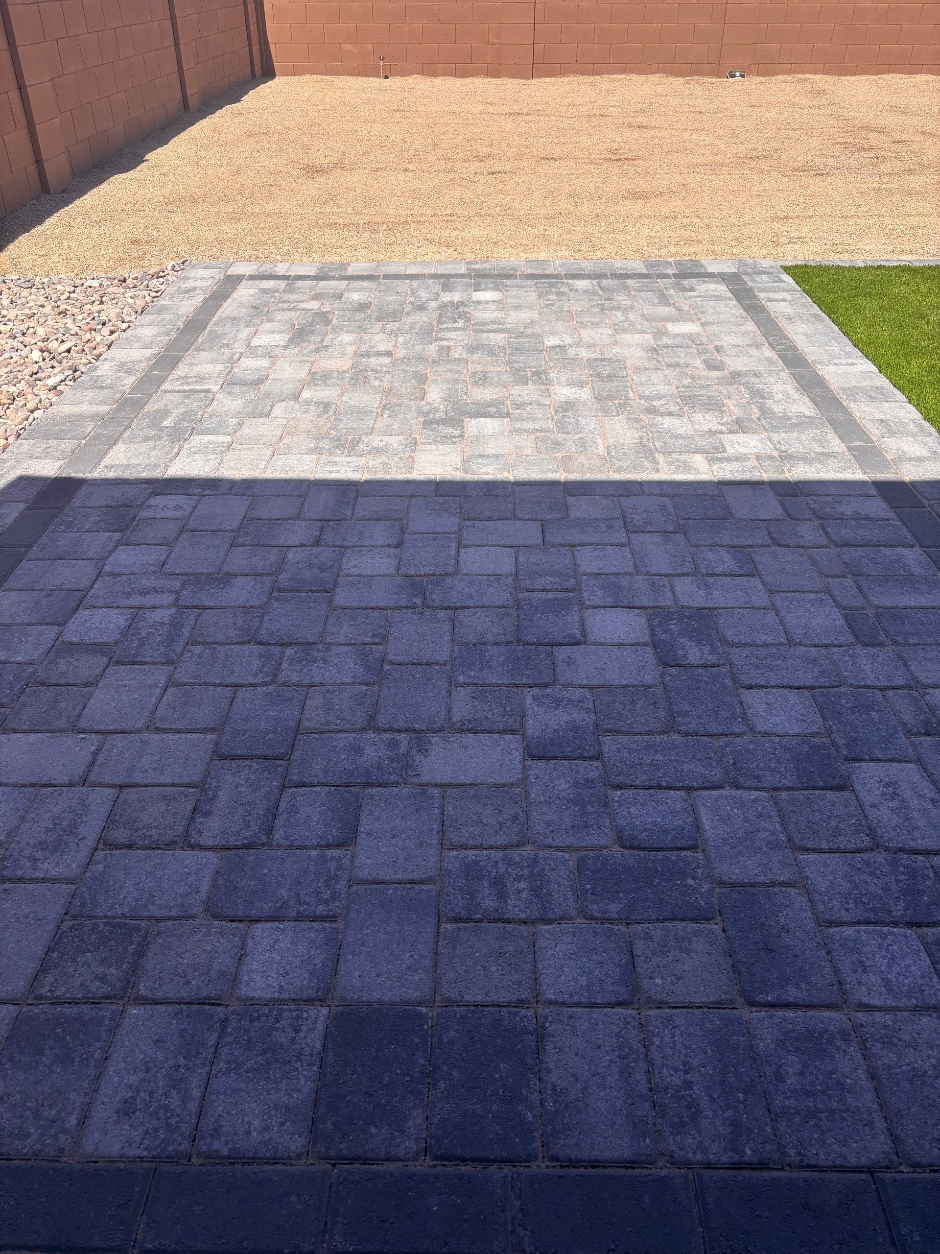 Patio with gray stone pavers, bordered by a lighter gray stone and beige gravel, with green grass to the side.
