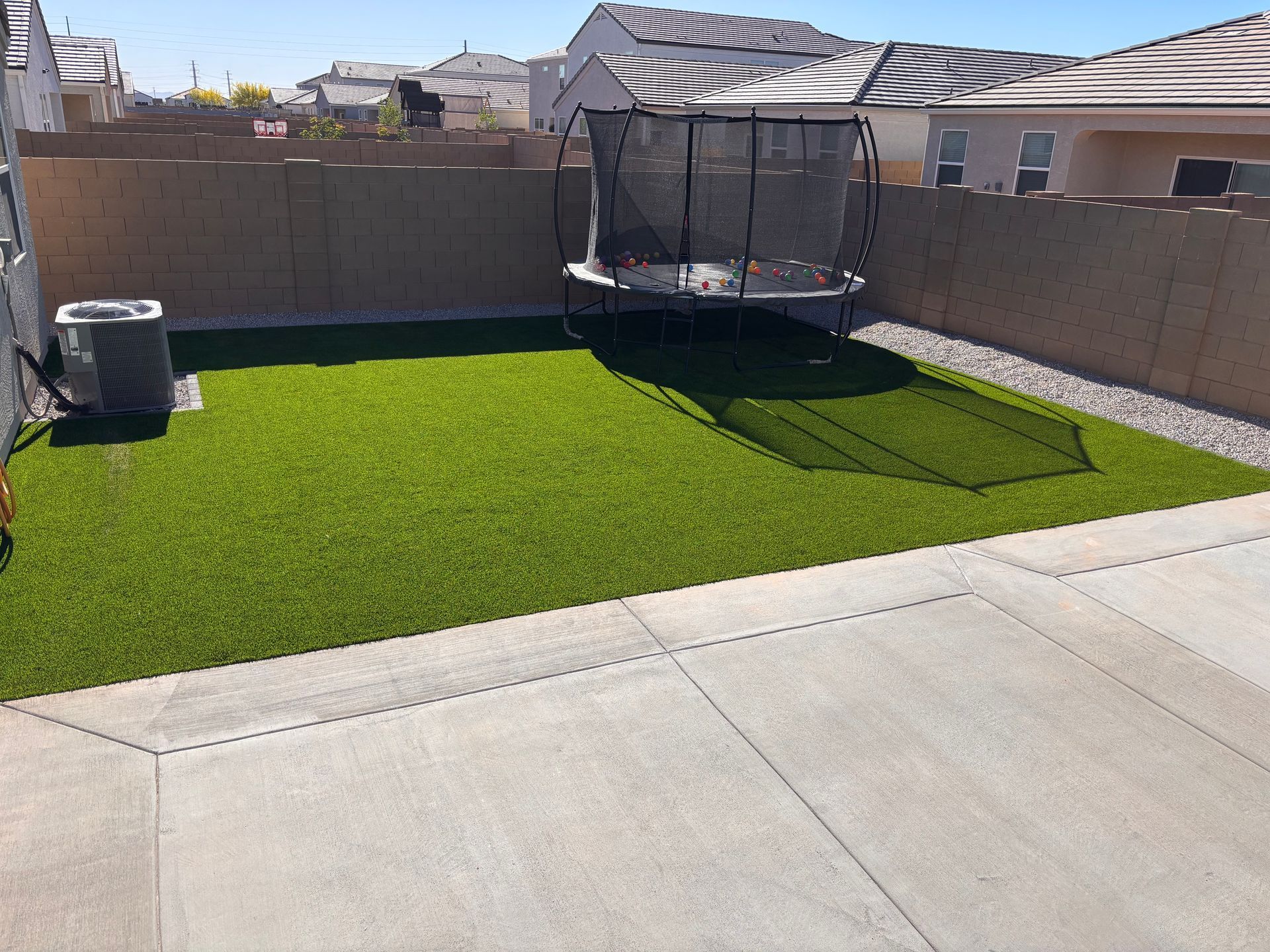 Backyard with artificial grass, trampoline, concrete patio, and air conditioning unit against a tan fence.