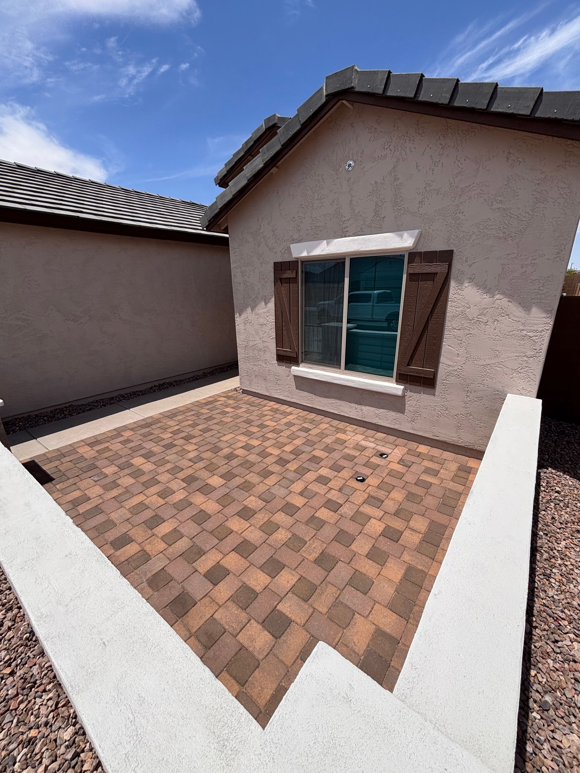 Small patio with brick pavers next to a tan stucco building with a window and brown shutters. Blue sky.