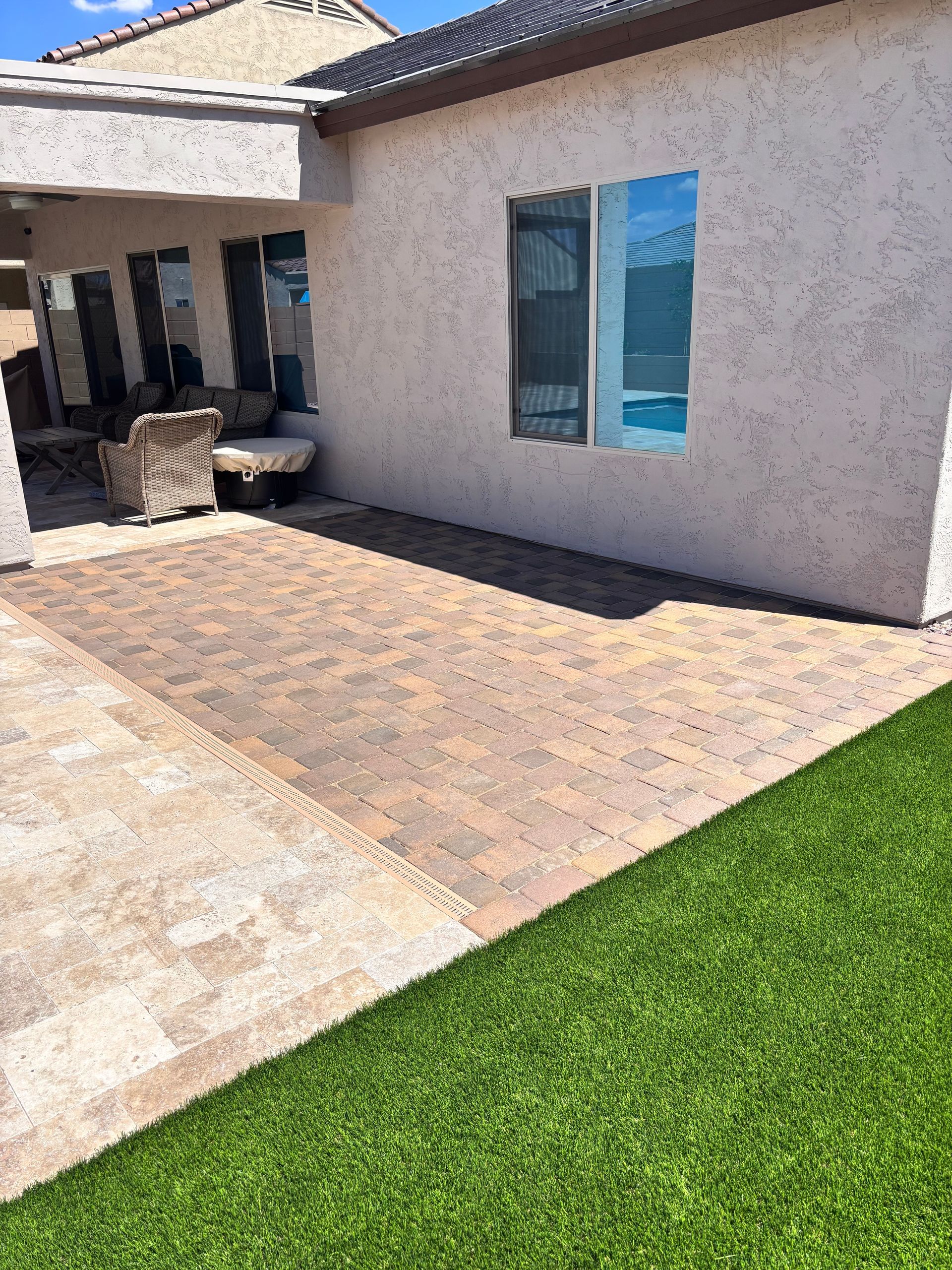 Patio with brick pavers next to green artificial turf, adjacent to a stucco building with a covered patio area.