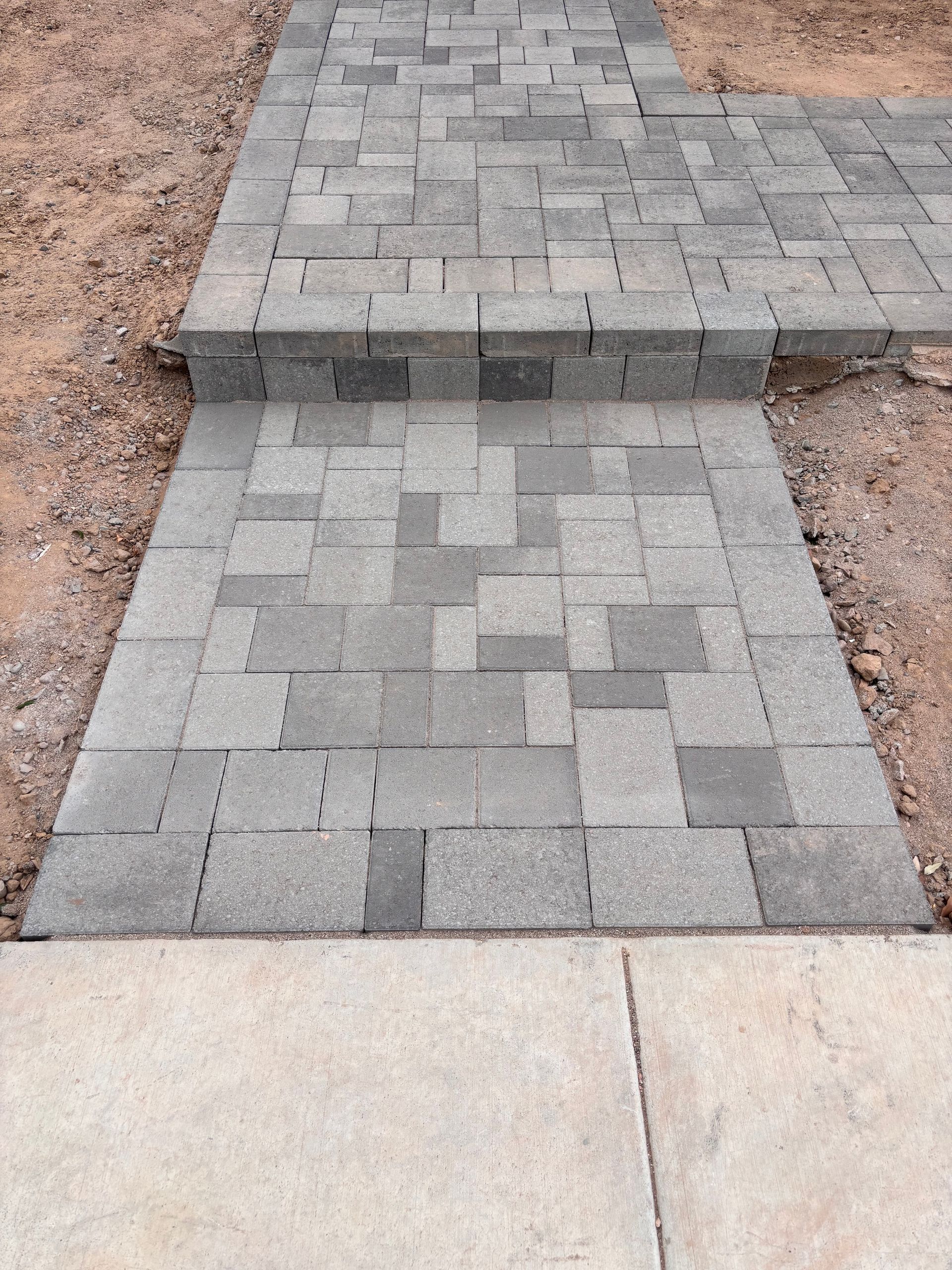 Brick pathway with a step, leading from a concrete sidewalk into a dirt area.