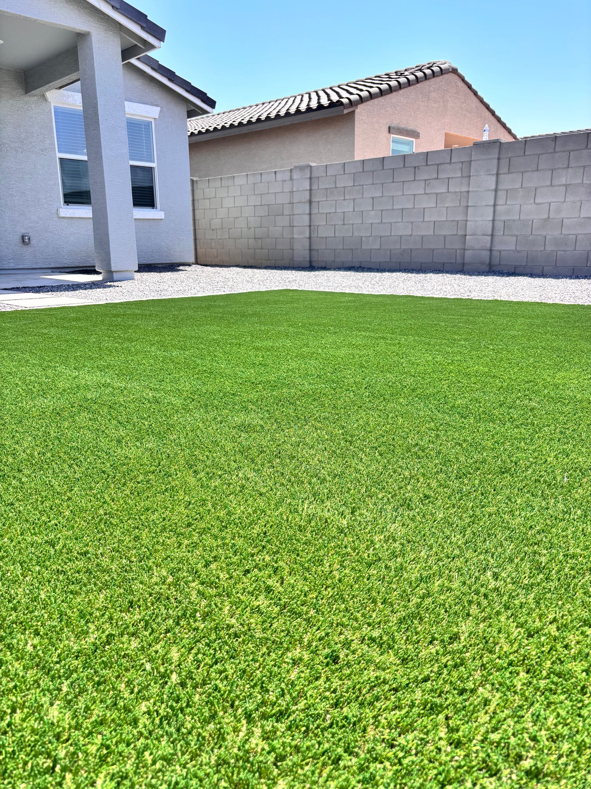 Green artificial grass in a backyard, with a house and block wall in the background under a blue sky.