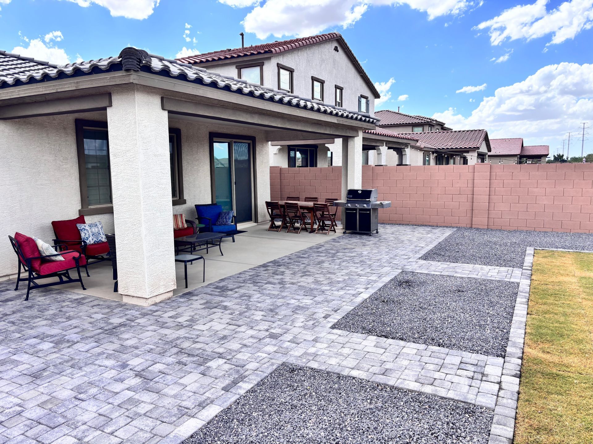 Patio with seating, dining table, and grill, paved with brick and gravel; house and wall in background.