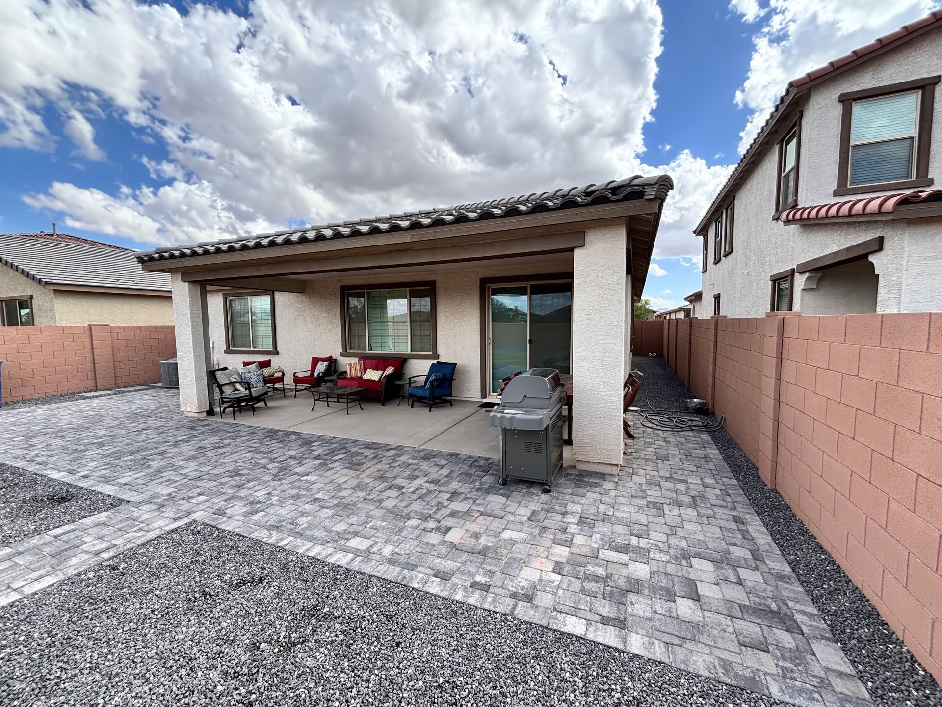 Backyard patio with paving stones, covered seating area, and brick wall under a cloudy sky.