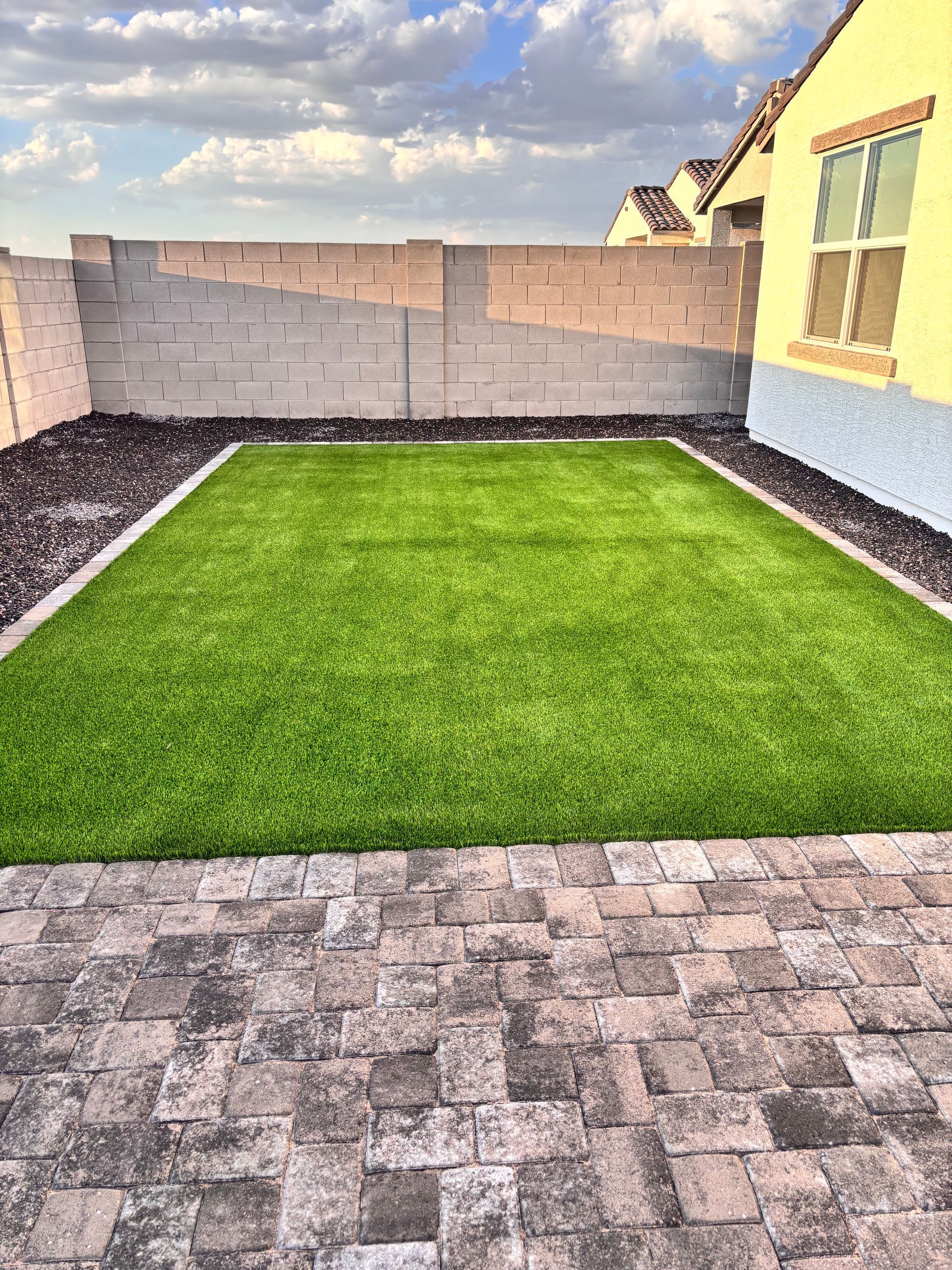 Backyard with brick patio, artificial grass, and beige walls under a partly cloudy sky.