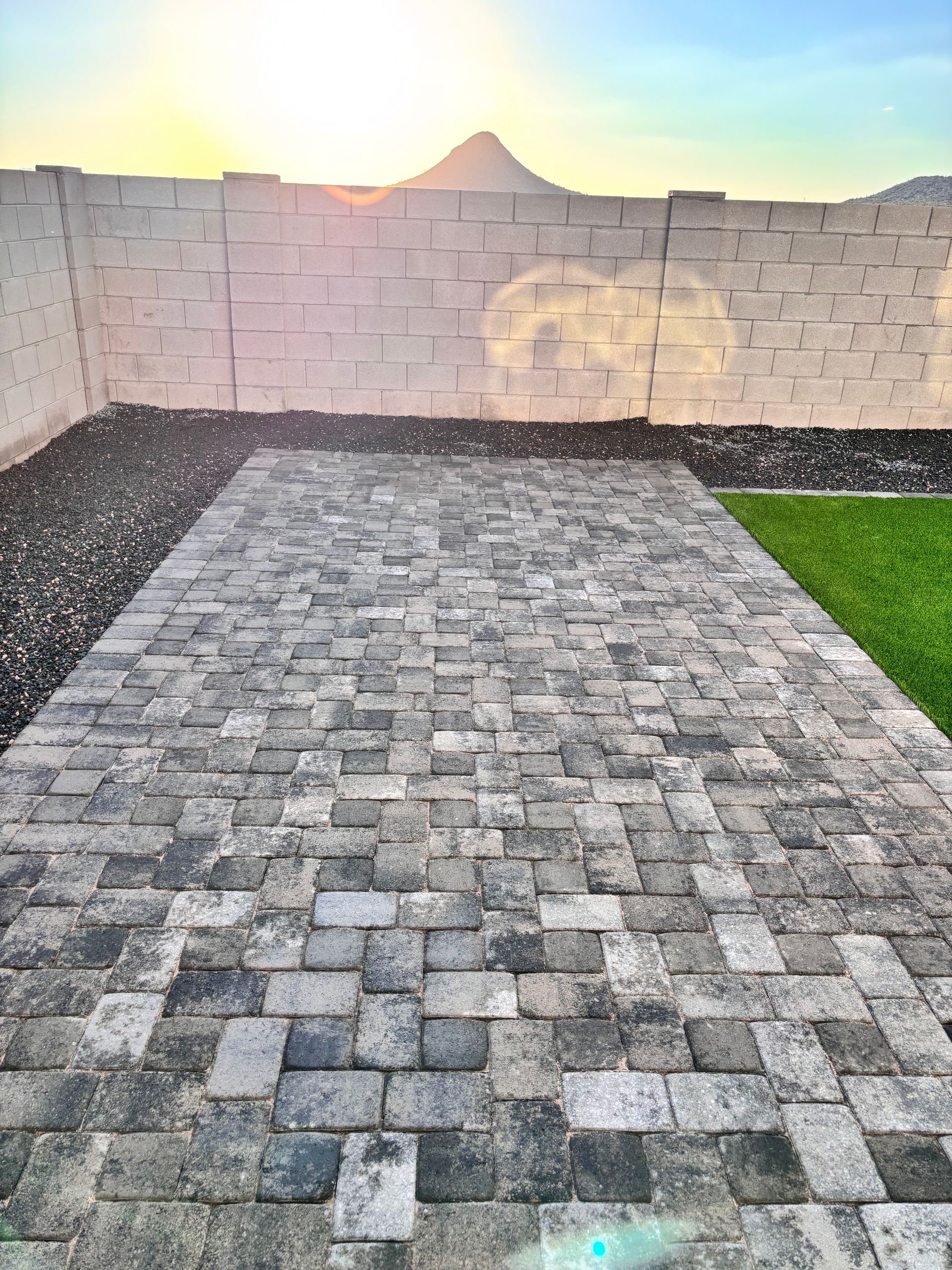 Stone patio leading to a wall with a mountain and setting sun in the background, next to green grass and black pebbles.