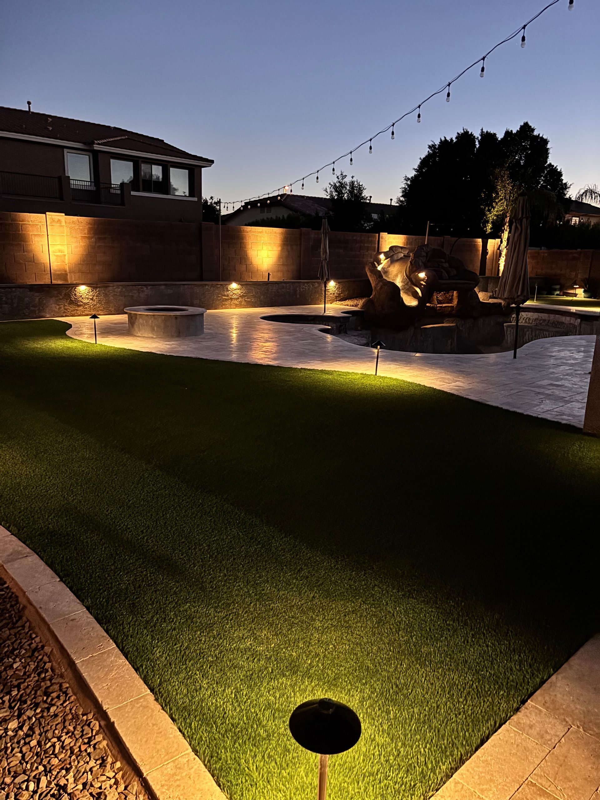 Backyard at dusk with string lights and illuminated landscaping. Green turf, paved areas, and a fire pit.