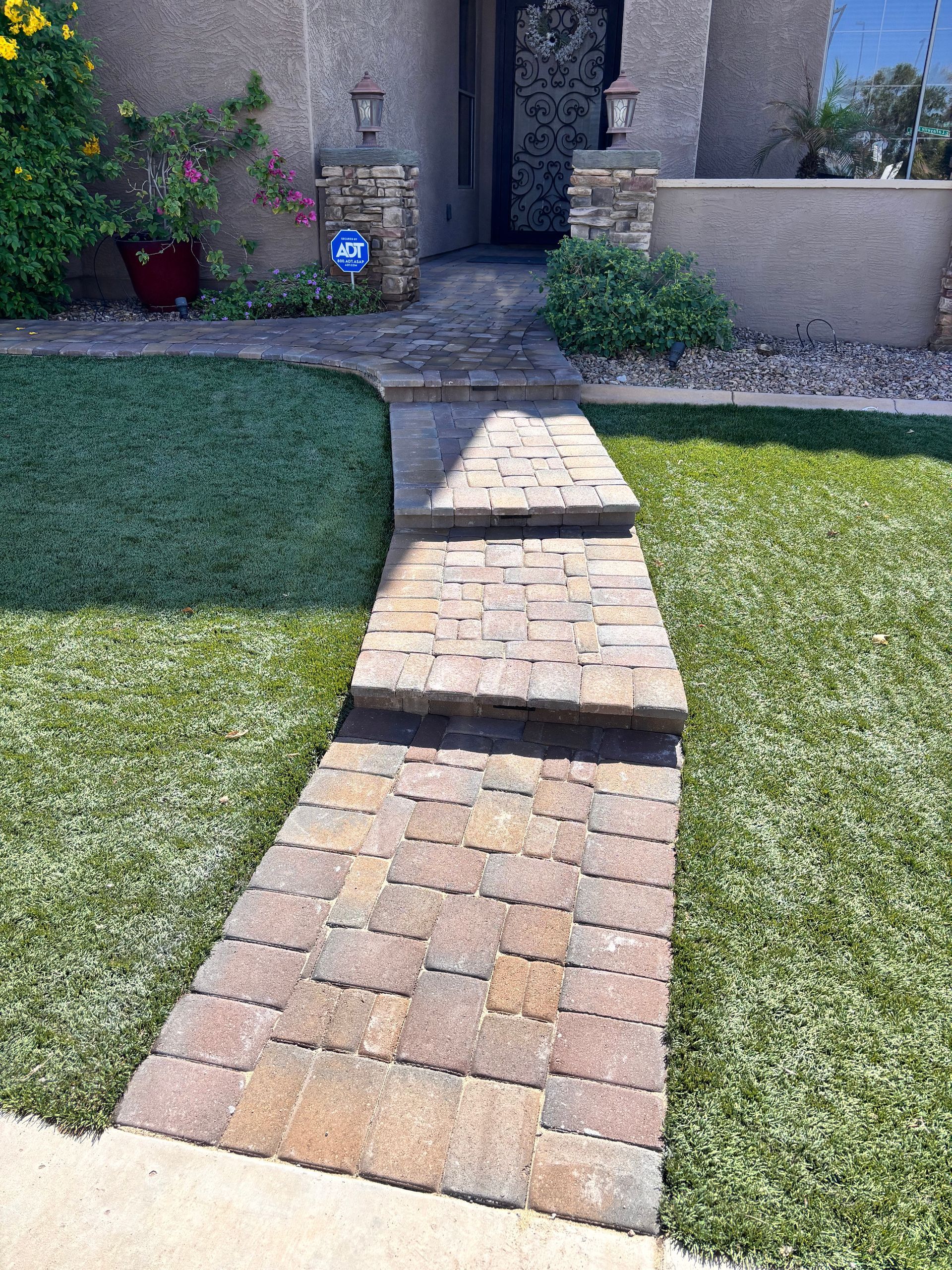 Brick pathway leading to a house entrance with steps, surrounded by green grass and plants.