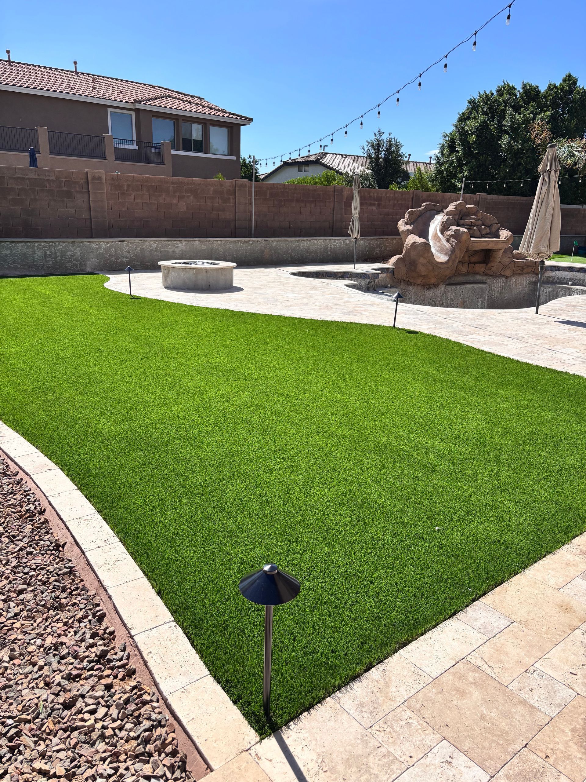 Artificial green turf in a backyard with a fire pit, decorative rocks, and a water feature under a sunny sky.