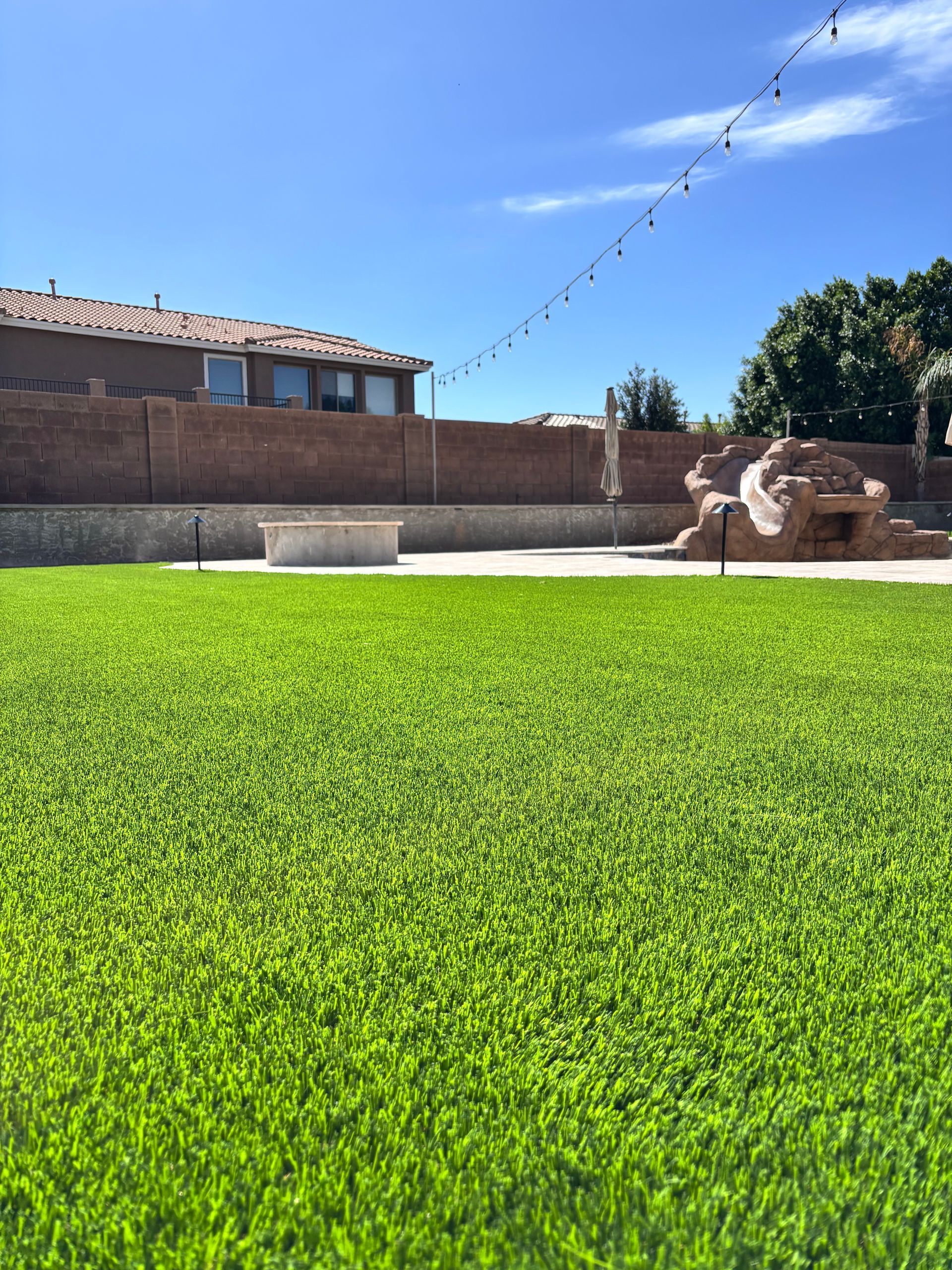 Green artificial lawn in backyard, bordering a pool. Red brick wall and house in background, clear blue sky.