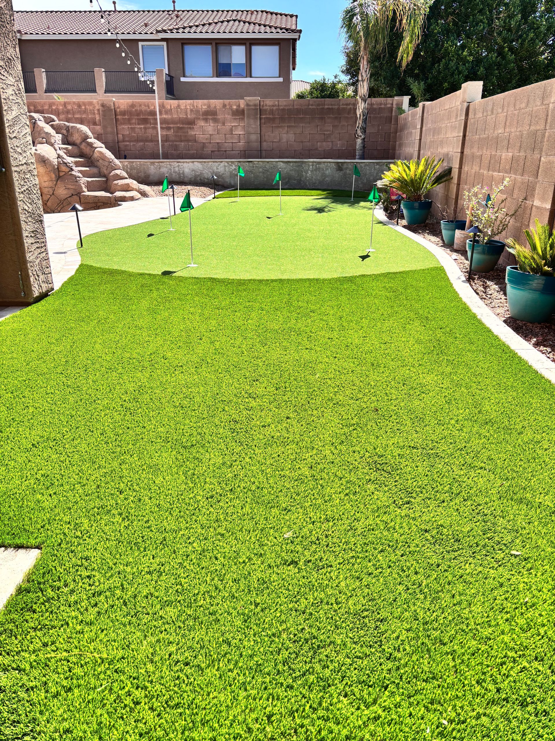 Backyard putting green with flags, surrounded by artificial turf and potted plants.