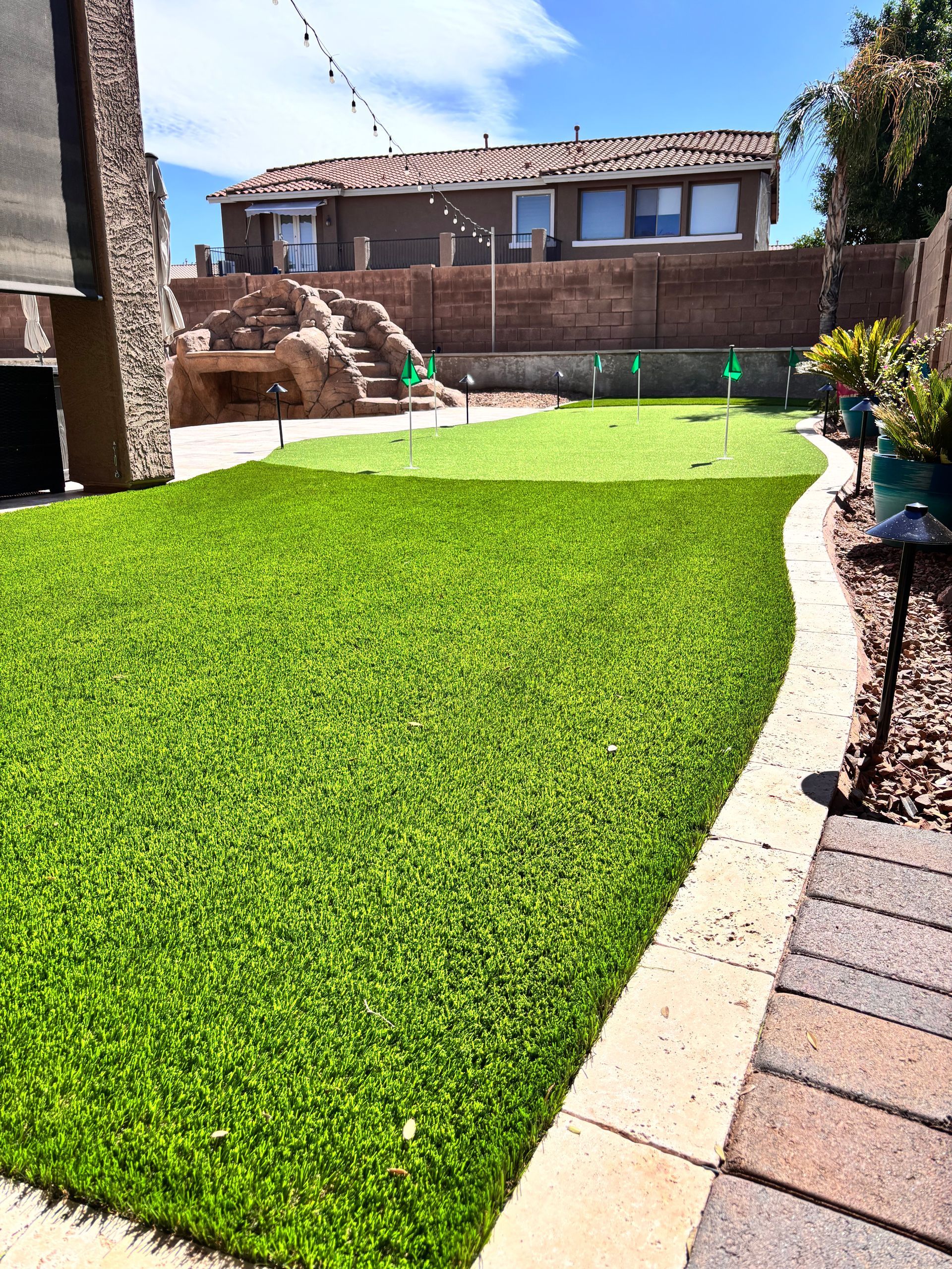 Artificial turf backyard with putting greens, bordering stone path, and a house in the background under a blue sky.