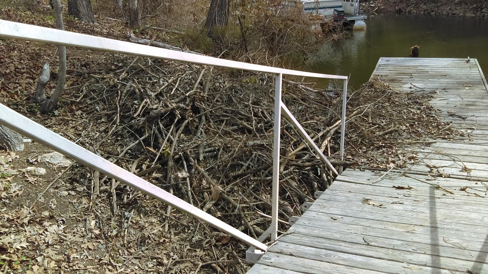 Wooden boardwalk along a muddy, debris-filled ditch with leaning railings in a rural area