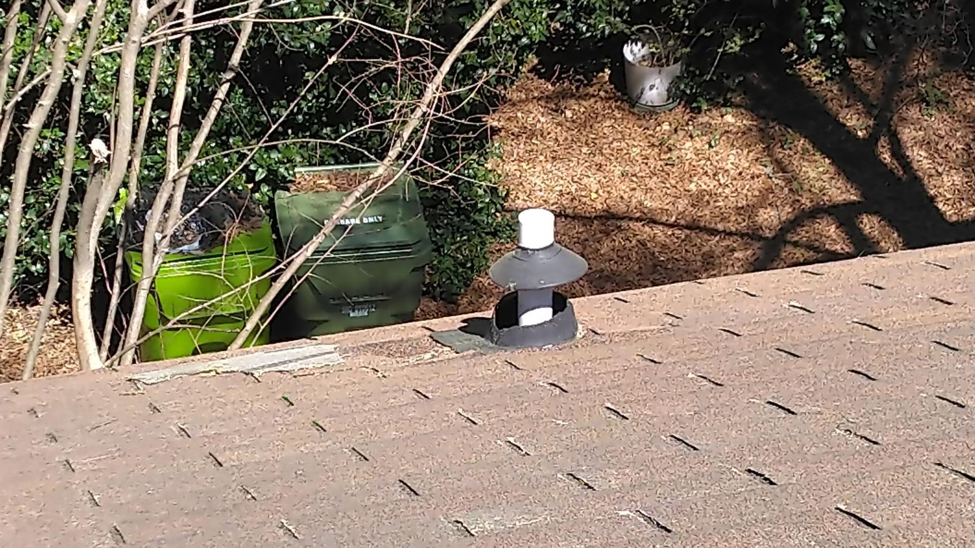 Roof edge with a small black-and-white vent cap near a leafy yard and mulch bed below.