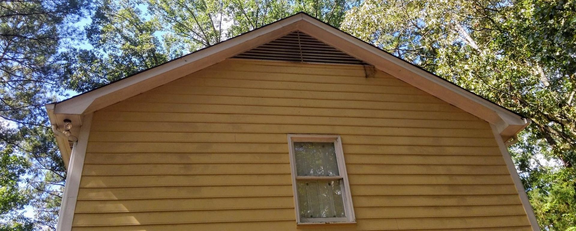 Yellow house gable with a small centered window, framed by trees and blue sky