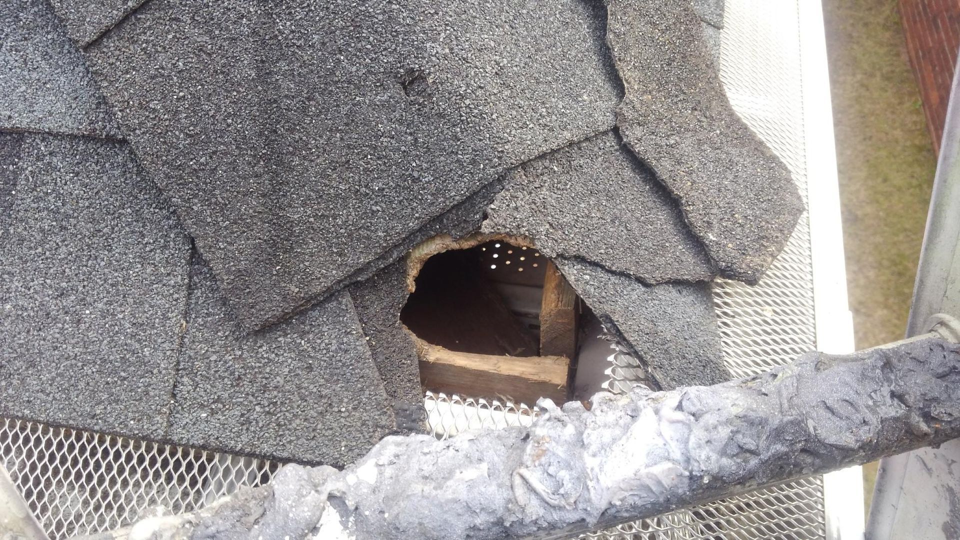 Close-up of a damaged shingled roof with a hole exposing wooden framing and debris.