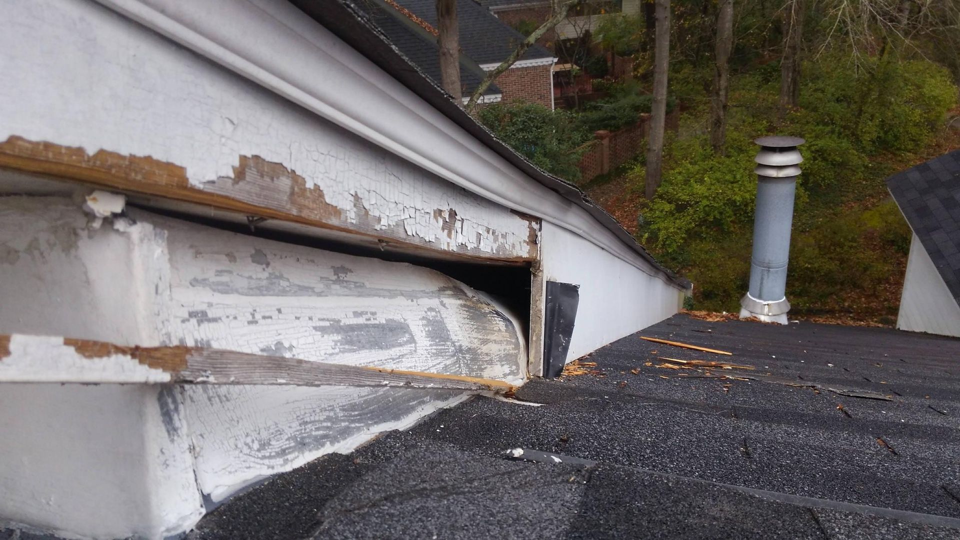 Damaged roof edge with peeling white siding and a black chimney vent on a shingled rooftop