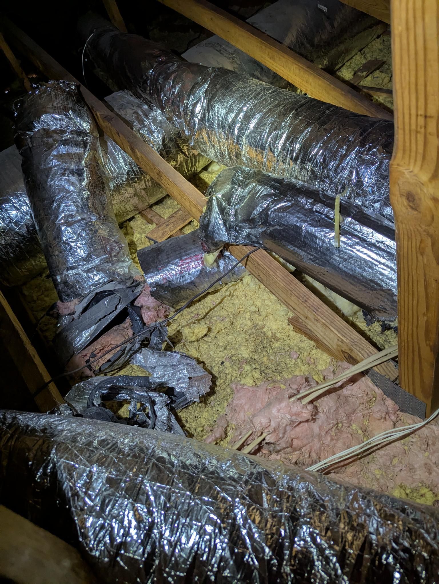Insulated HVAC ducts in an unfinished attic with exposed wooden rafters and pink insulation.