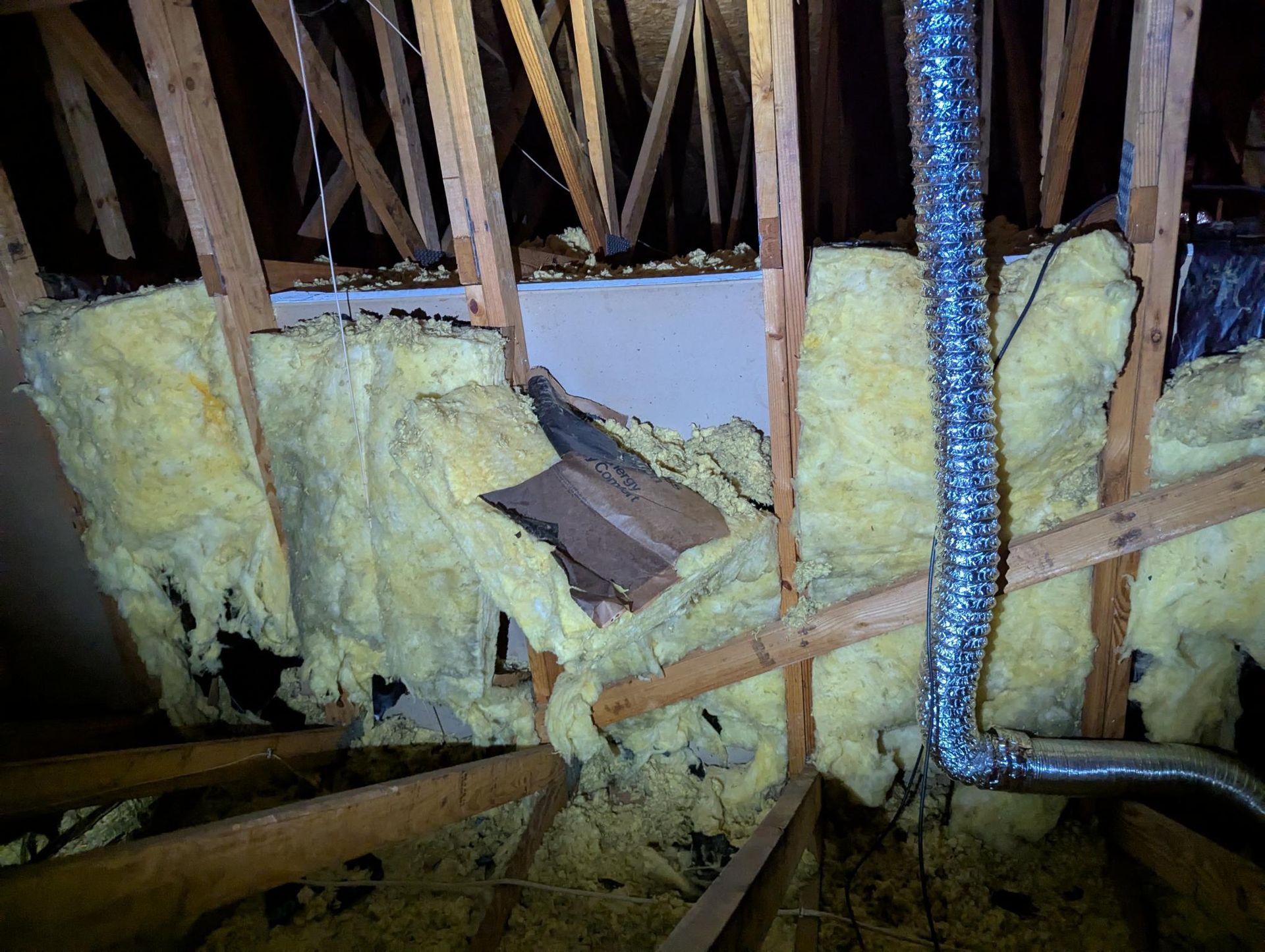 Attic with exposed wooden framing, yellow insulation, and a silver flexible duct hose