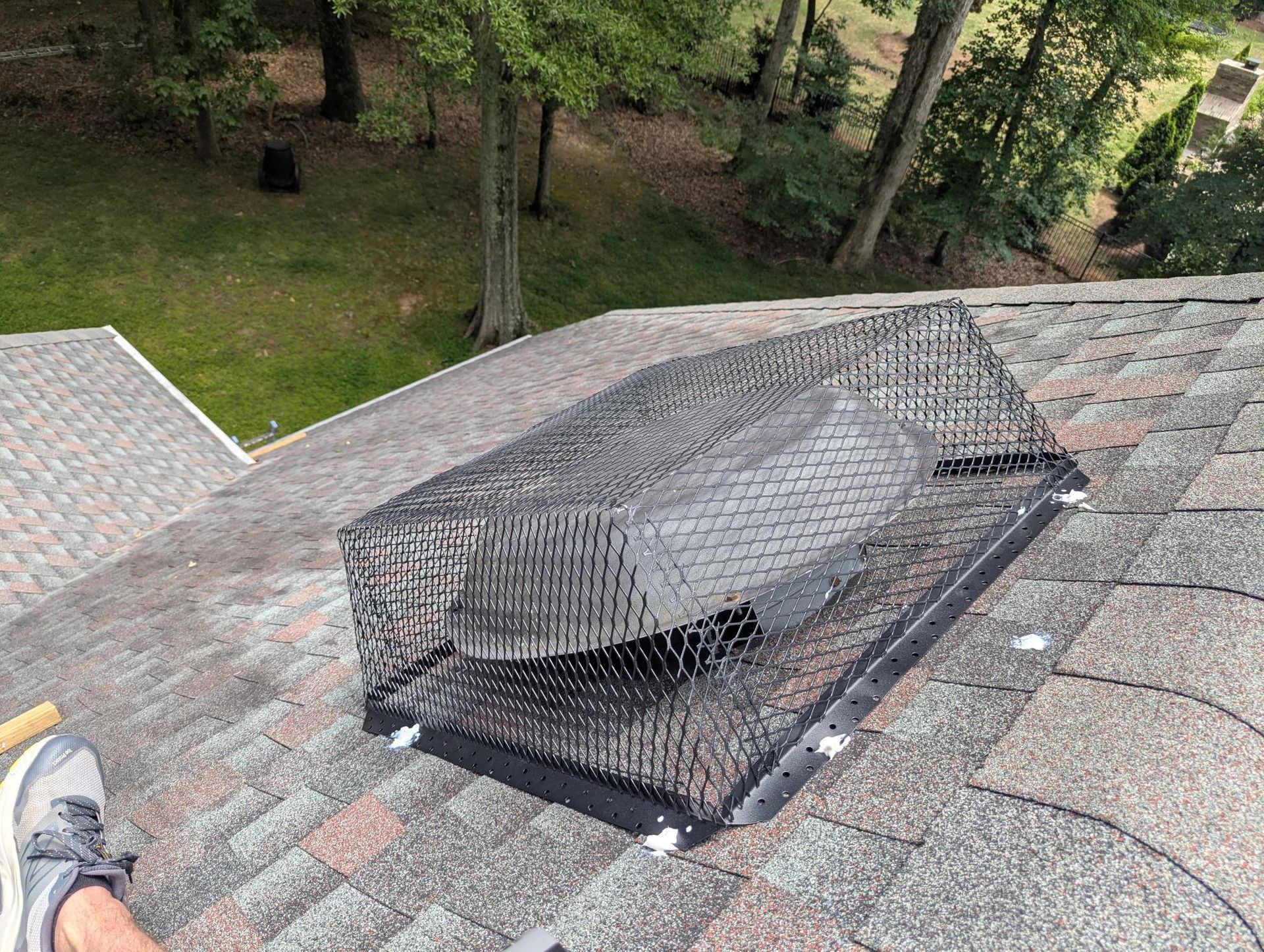 Black chimney vent capped with wire mesh on a shingled roof, with trees in the background