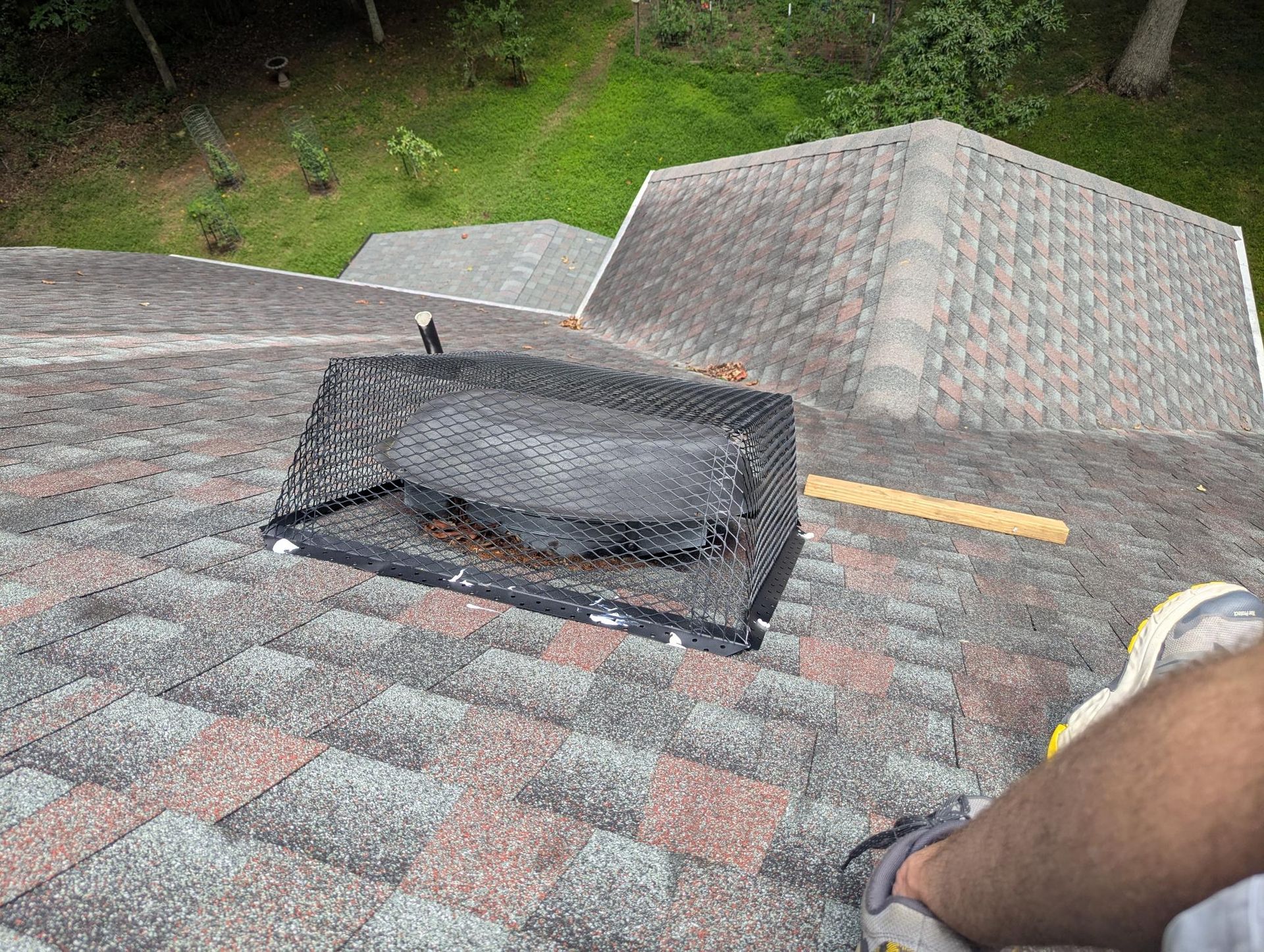 Rooftop with a damaged black vent cover on gray shingles, viewed from above near a wooded area.