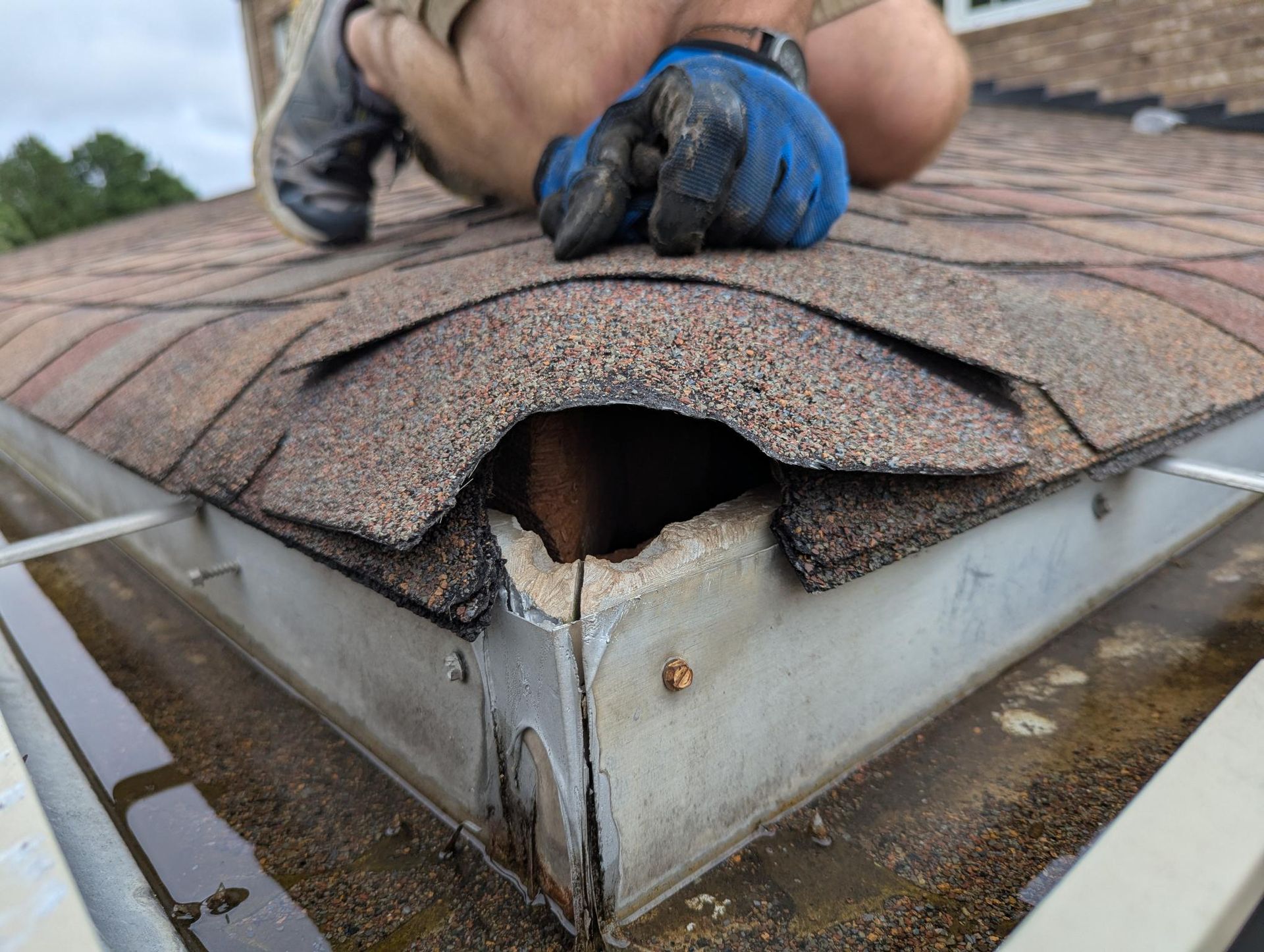 Damaged shingle roof with a large hole and a worker’s gloved hands inspecting it