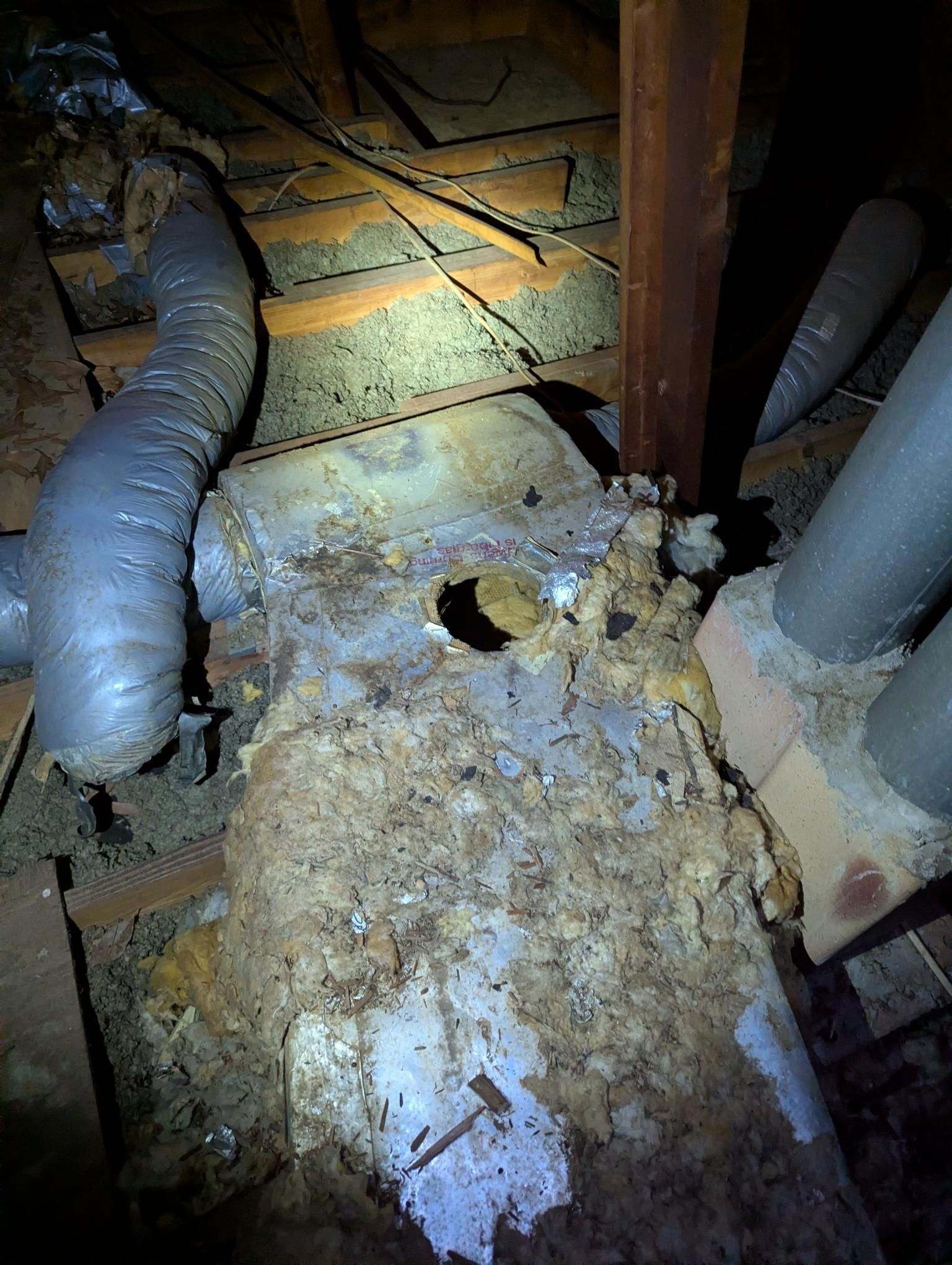Damaged attic ductwork with a hole in insulation and debris scattered on the floorboards