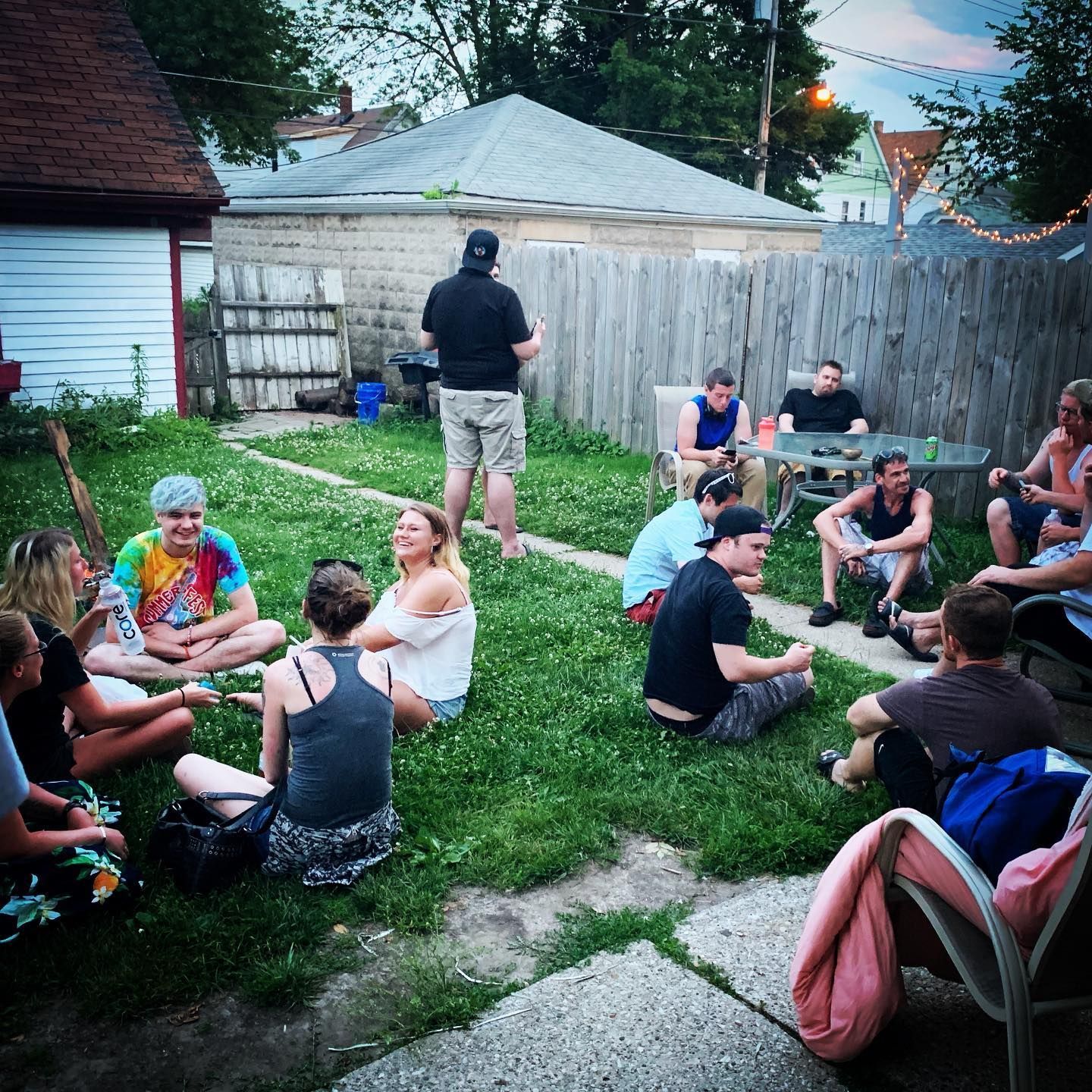 People sitting and standing in a backyard, socializing. Wooden fence, lawn, and various houses in background.