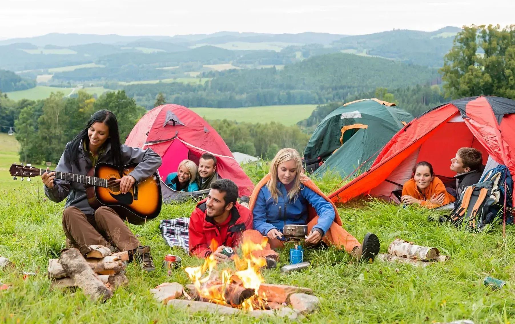 Campers sit and play guitar around a campfire beside colorful tents in a grassy field with hills beyond.