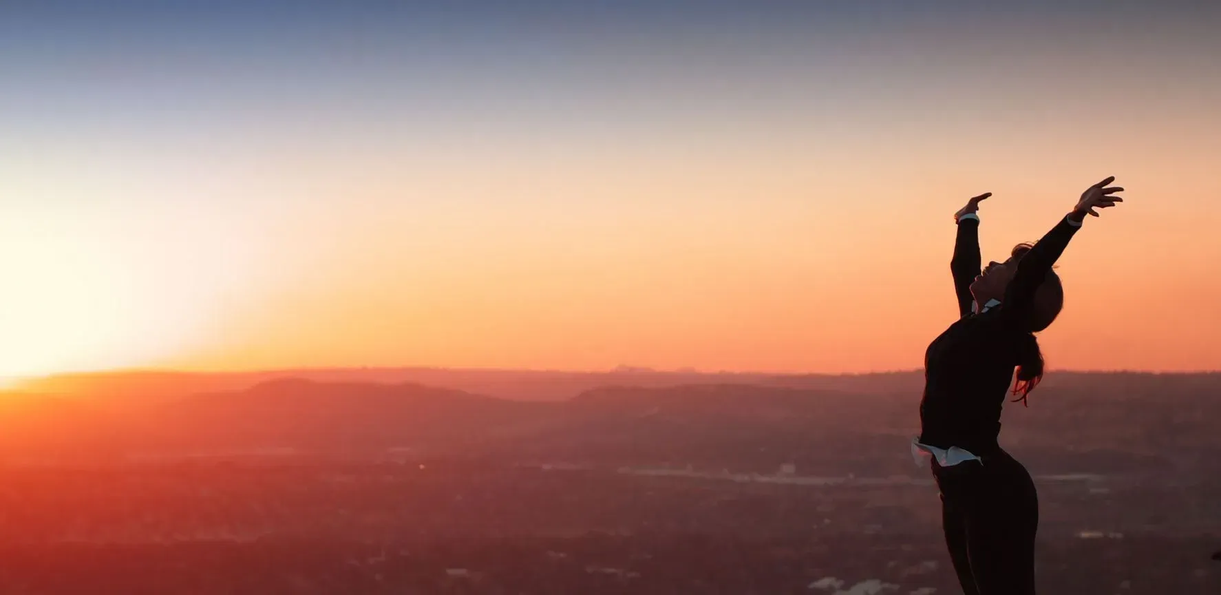 Silhouette of a person with arms raised, facing a sunset over mountains.