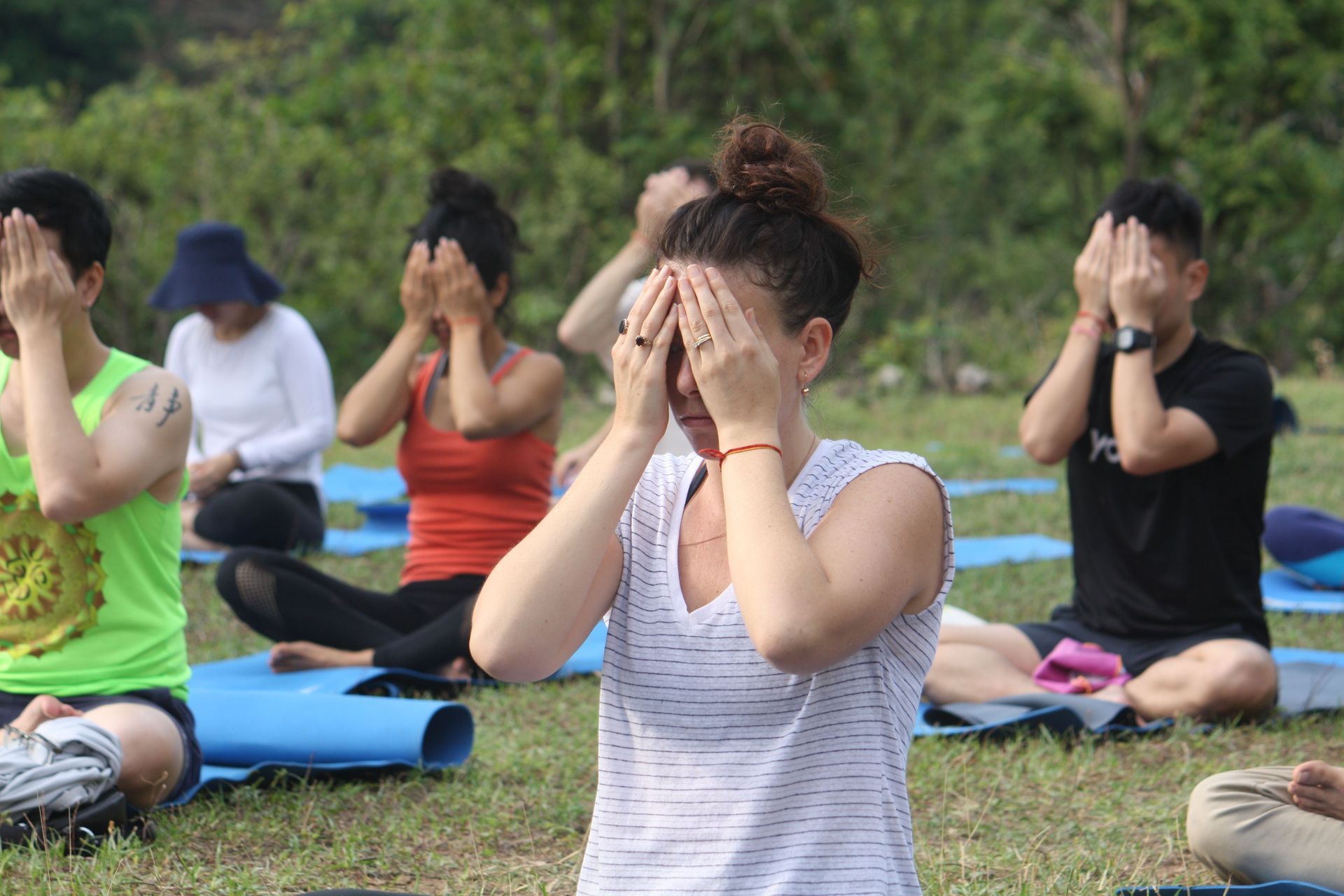 Group doing seated yoga on mats outdoors, covering their faces with both hands.