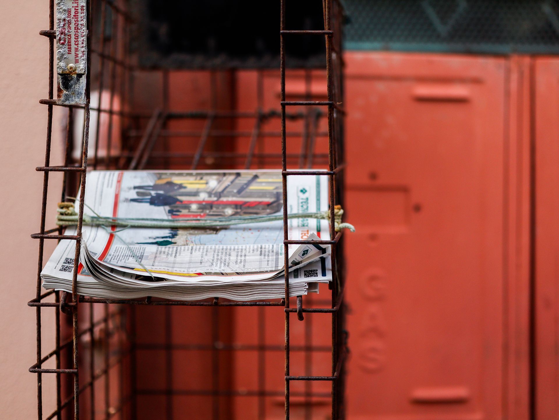 Ein Zeitungsstapel in einem verrosteten Metallkäfig vor einer verblassten roten Wand.Fotograf Tübingen. Fotograf Reutlingen.