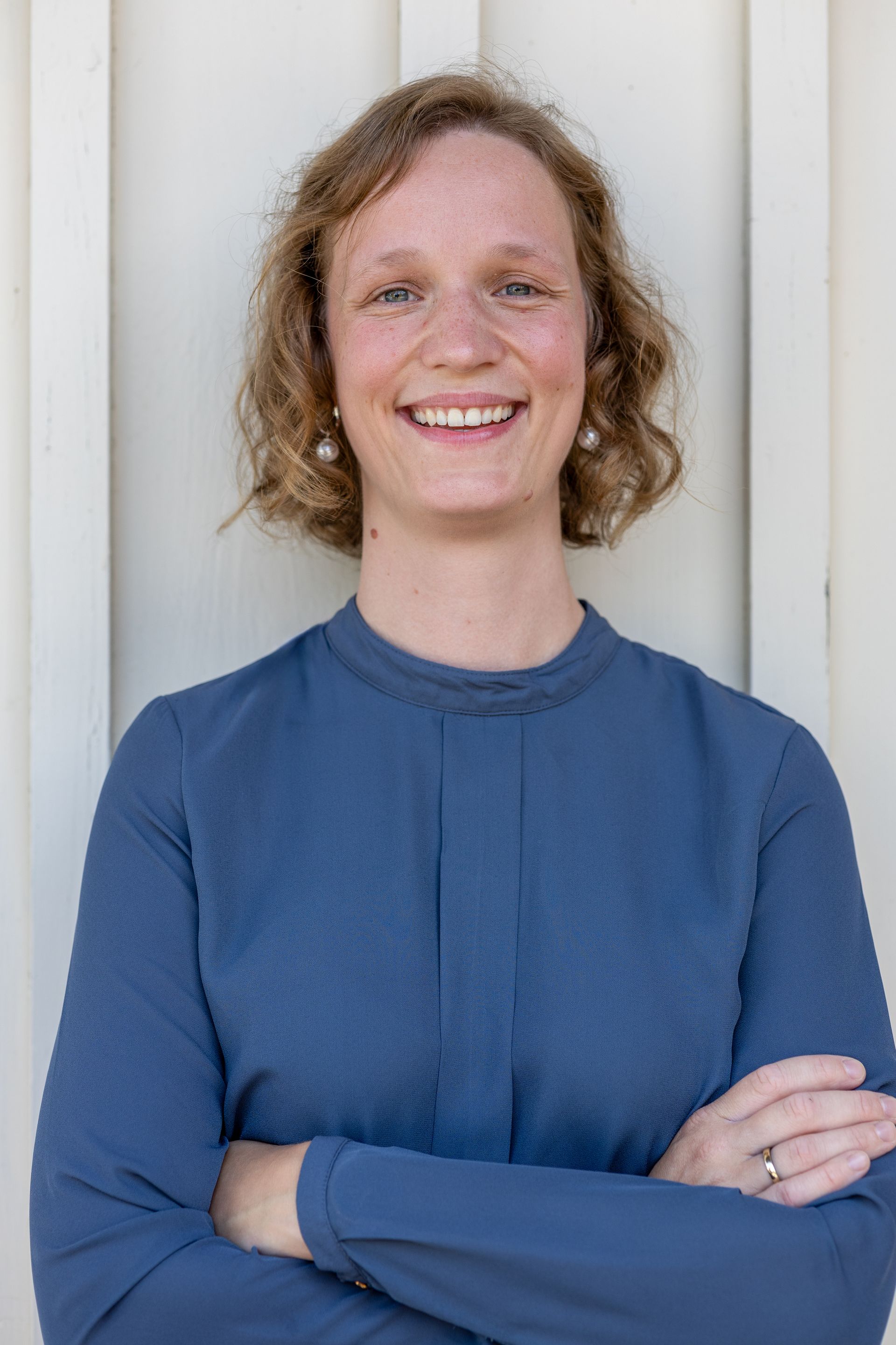Frau in blauem Oberteil, lächelnd mit verschränkten Armen, lehnt an weißer Holzwand.Fotograf Tübingen. Fotograf Reutlingen.