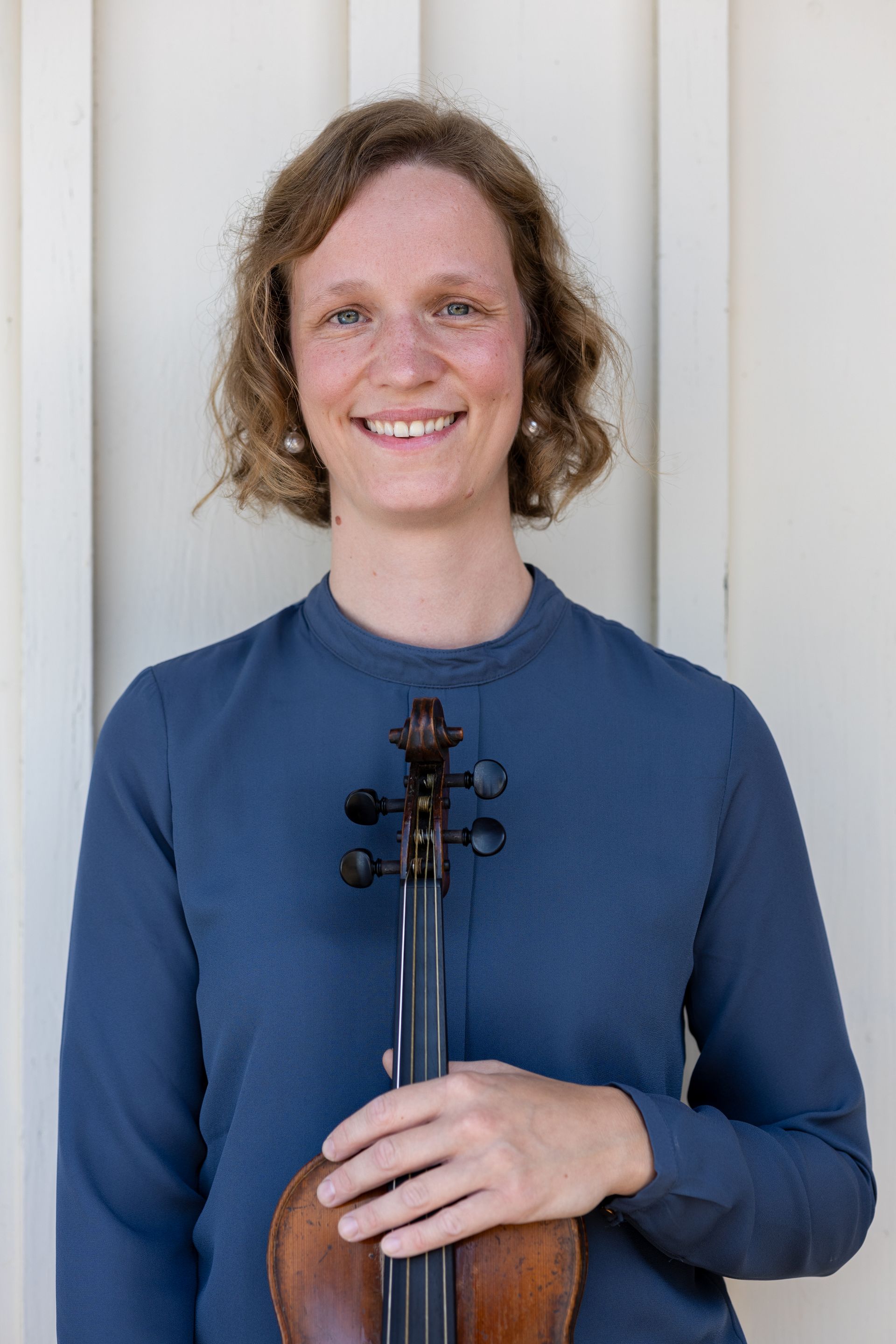 Frau in blauem Oberteil, die eine Geige hält, lächelt und lehnt an einer weißen Wand.Fotograf Tübingen. Fotograf Reutlingen.