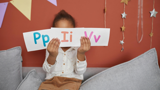 A little girl is sitting on a couch holding a sign that says pp ii vv.