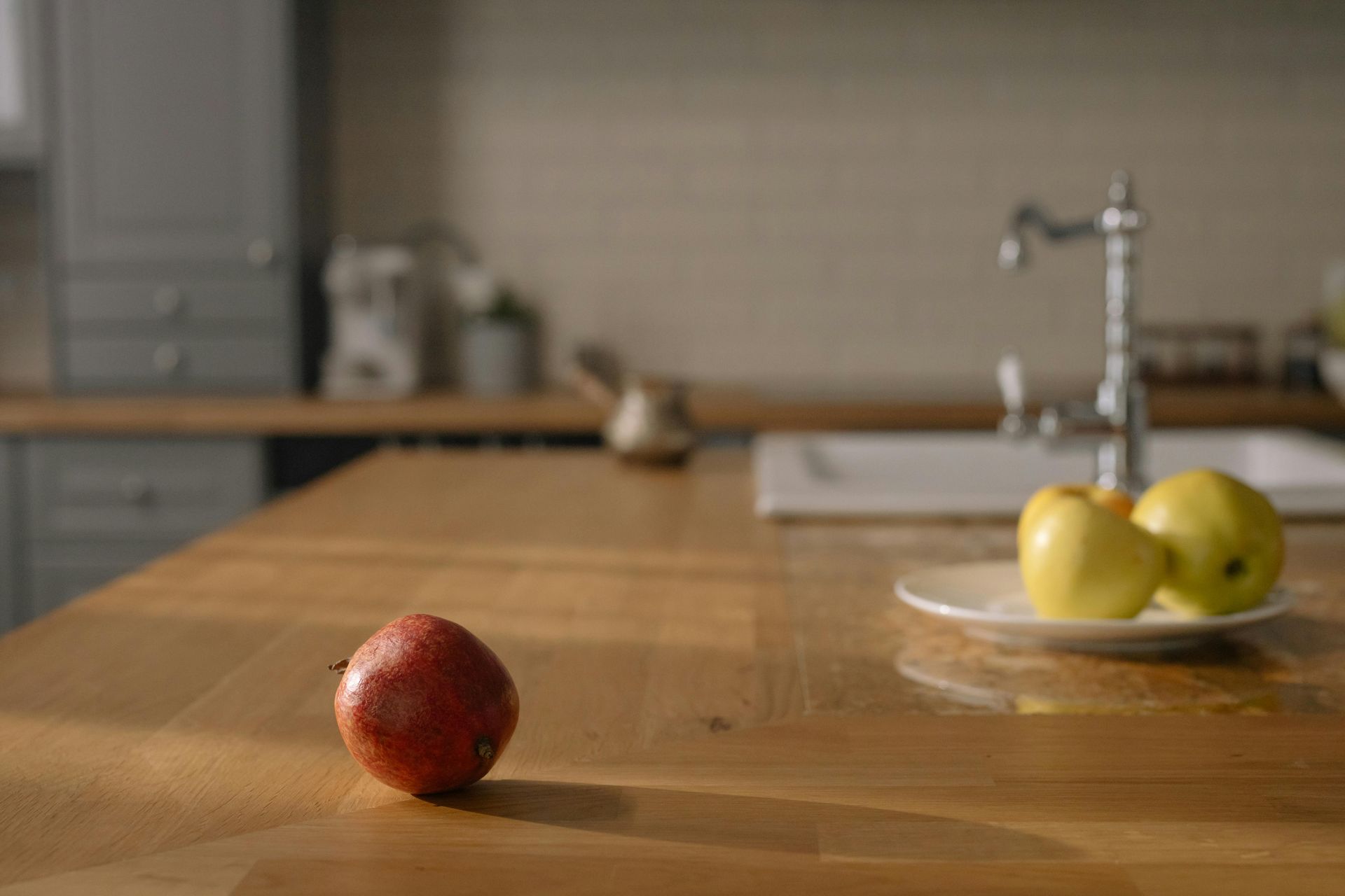 Red apple on a wooden countertop with two green apples near a sink in a kitchen.