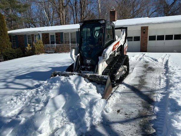 A white Bobcat compact track loader pushing snow off a residential driveway on a sunny winter day.