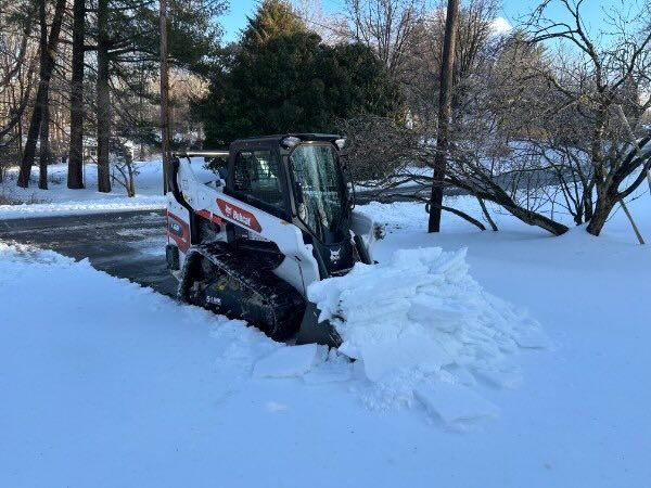 A white Bobcat skid-steer loader clearing heavy snow from a residential driveway on a sunny, winter day.