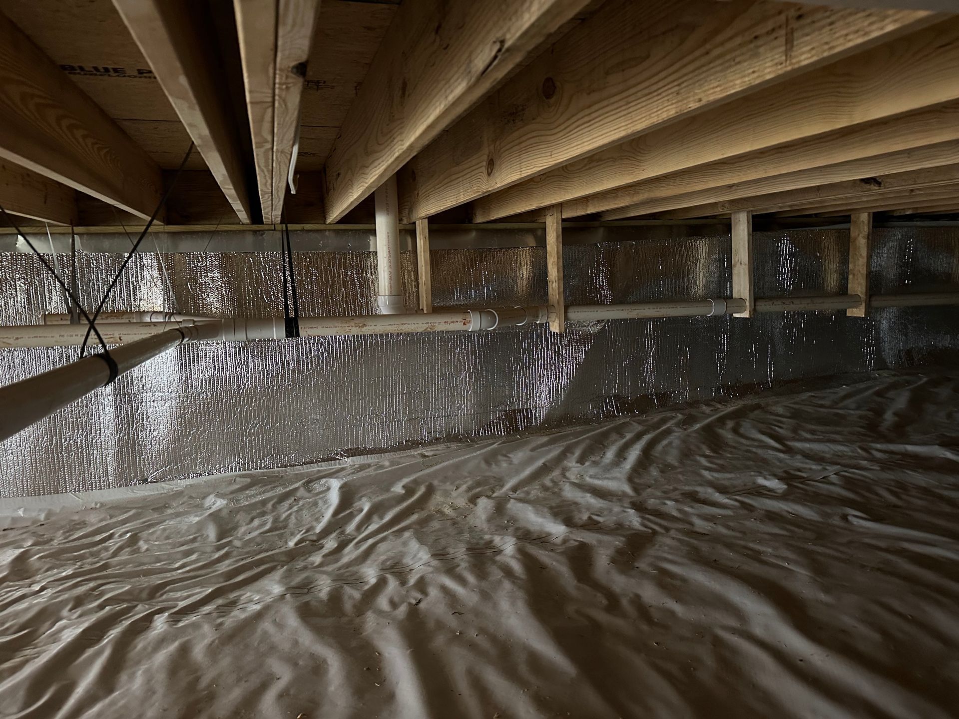 Crawlspace with insulation, vapor barrier, and wooden beams. Silver insulation reflects light.