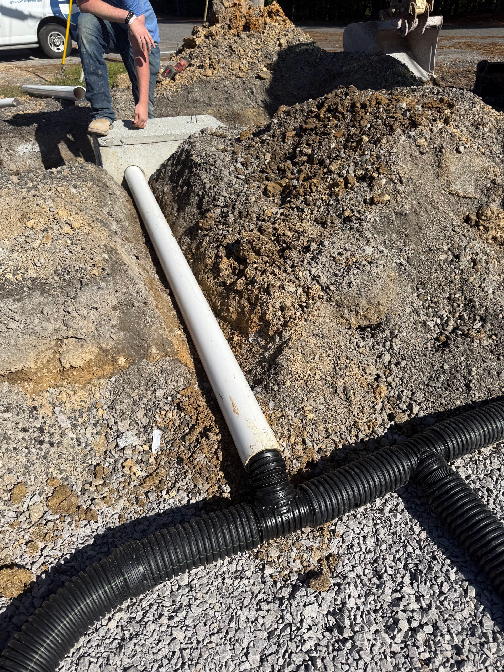 Construction site: white pipe connected to black corrugated drainage pipes in a gravel bed, dirt pile in background.