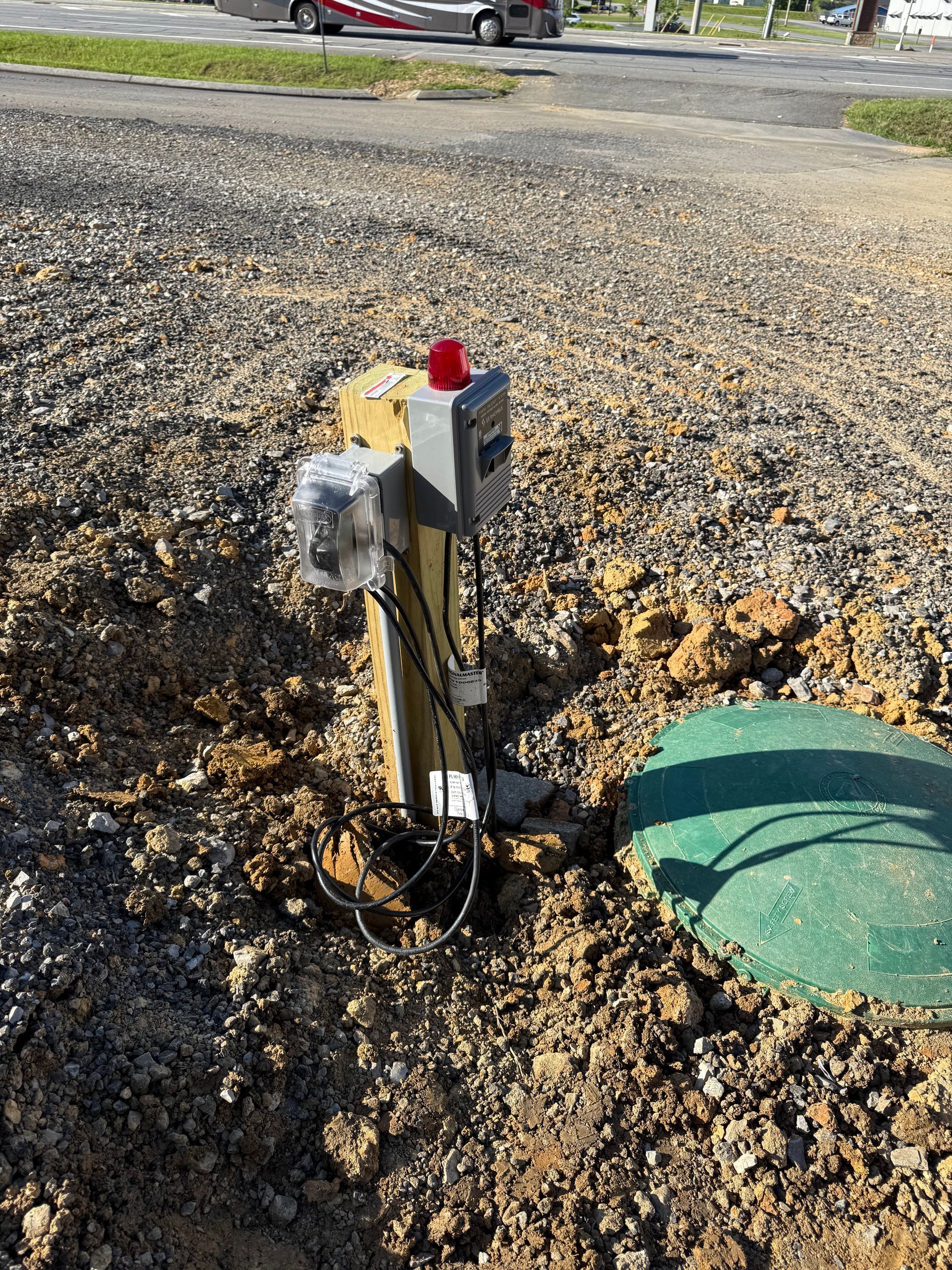 A green septic tank lid with a control box and alarm mounted on a post in gravel and dirt.